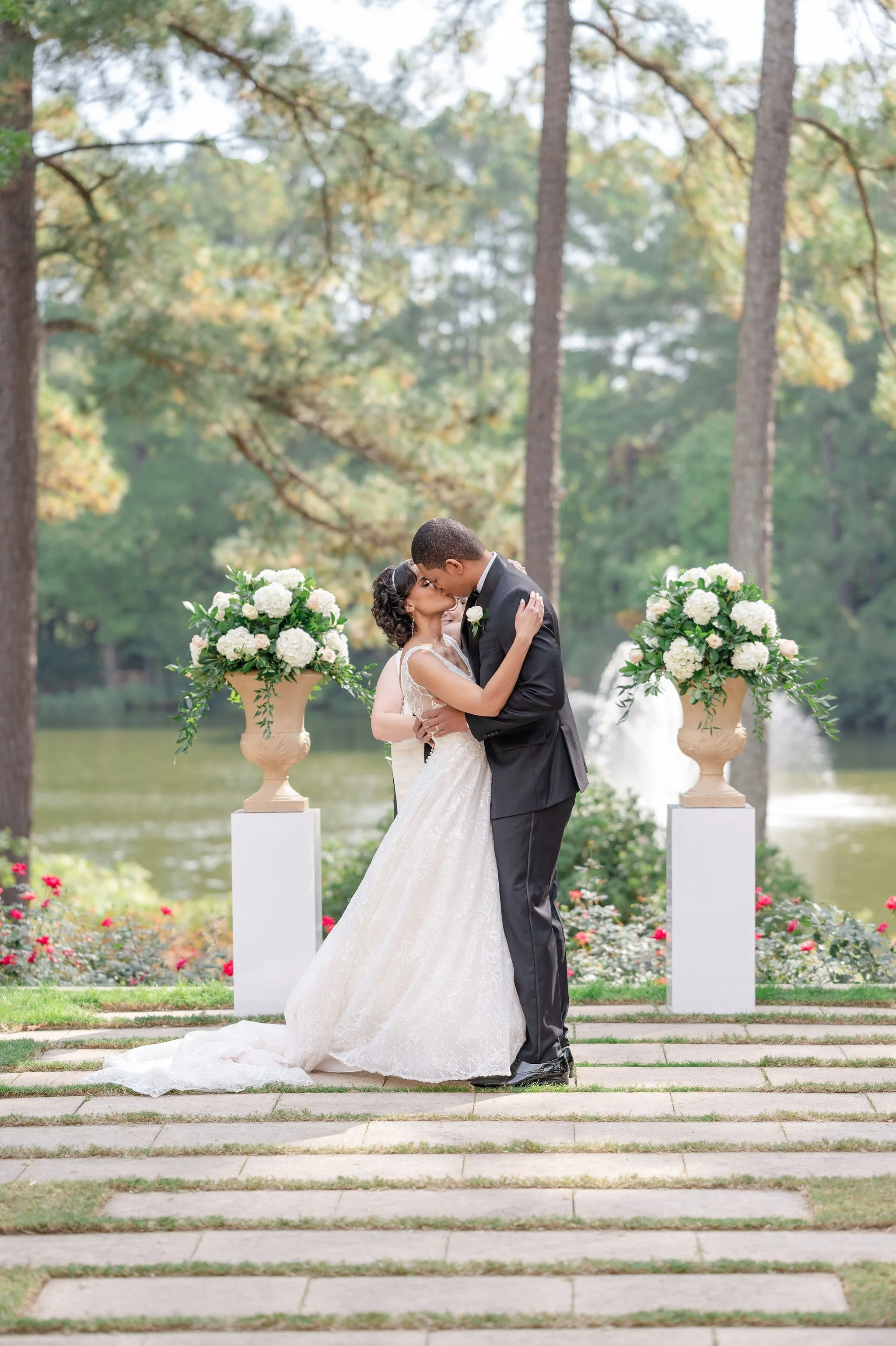A newlywed couple sharing a kiss and embracing outdoors near a lake, with trees and floral arrangements in the background.