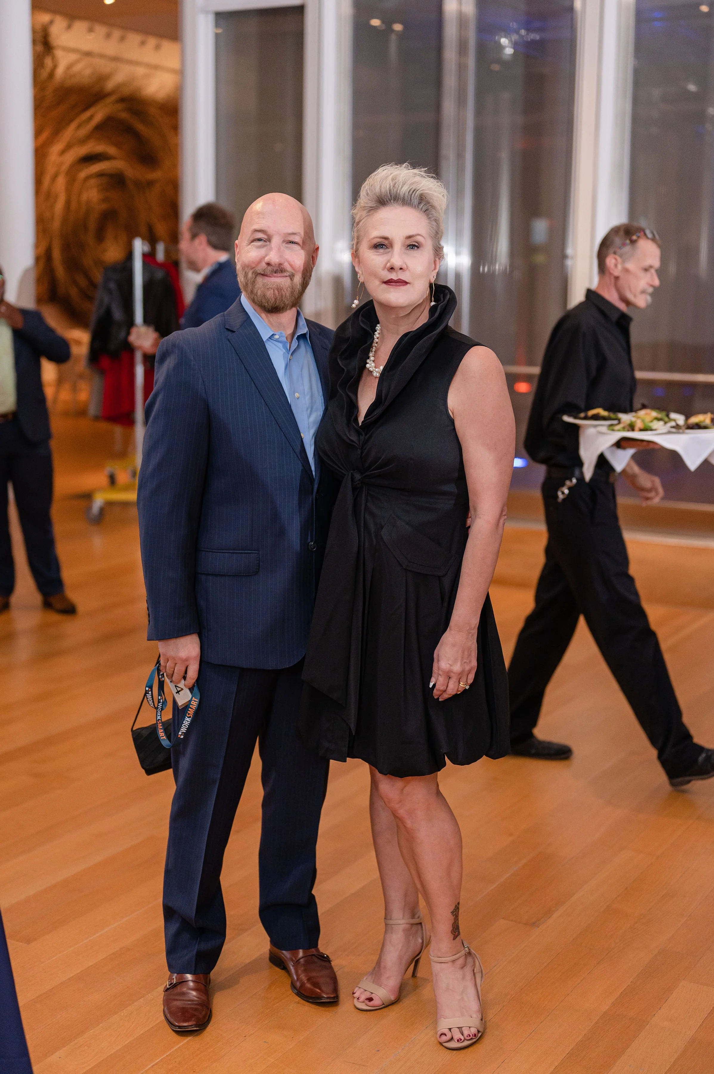A man and a woman dressed in formal attire standing in a well-lit indoor event space, with other people in the background including a waiter carrying plates of food.