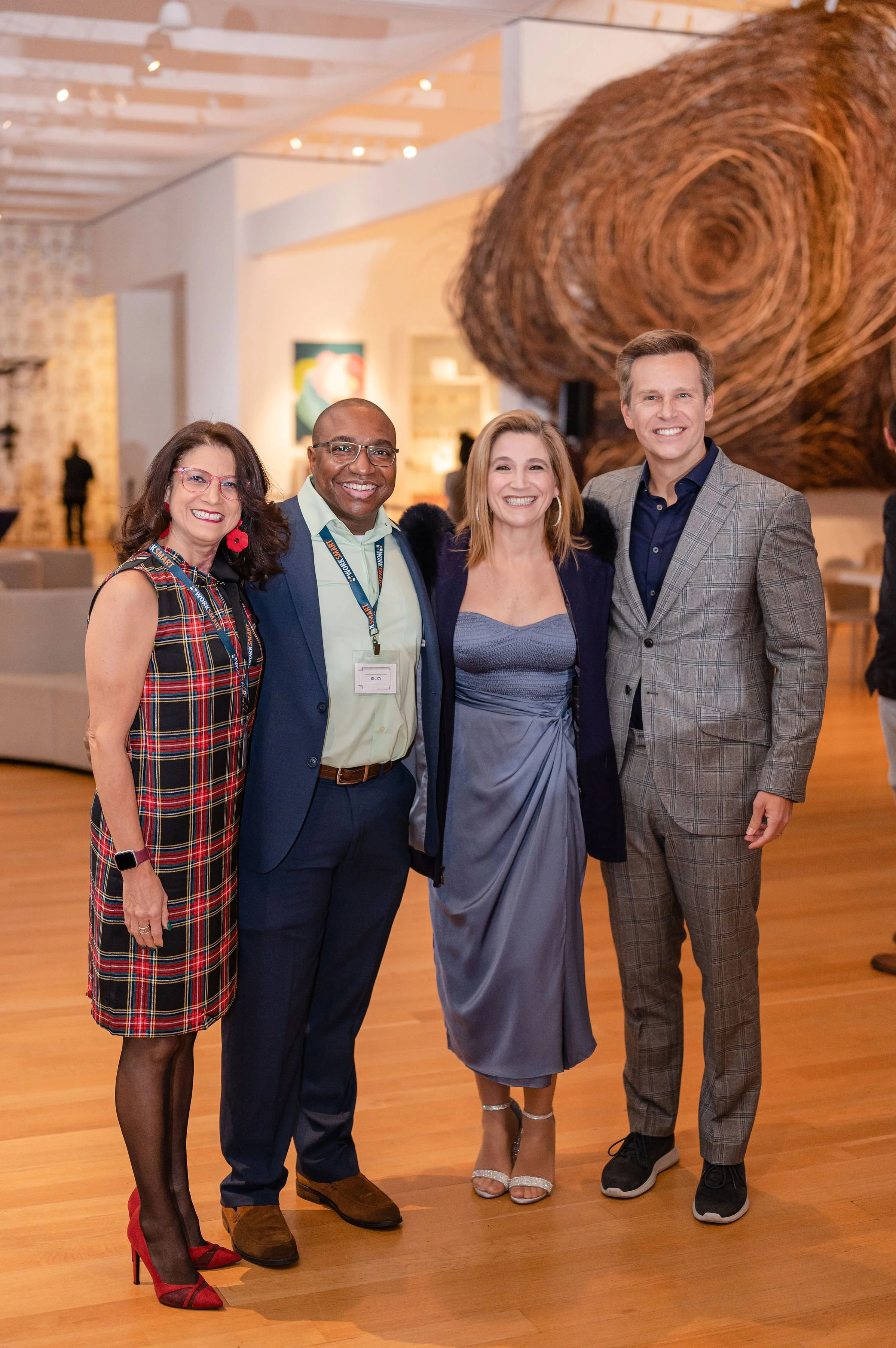Group of four people smiling and posing for a photo at an art gallery or museum, with colorful artwork on the walls and large wooden sculpture in the background.