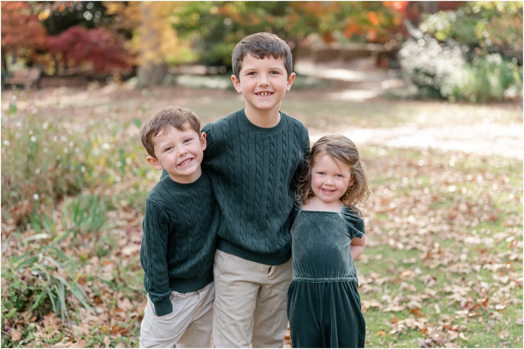 Three children standing outdoors in fall, smiling, with fallen leaves on the ground and trees with autumn foliage in the background.