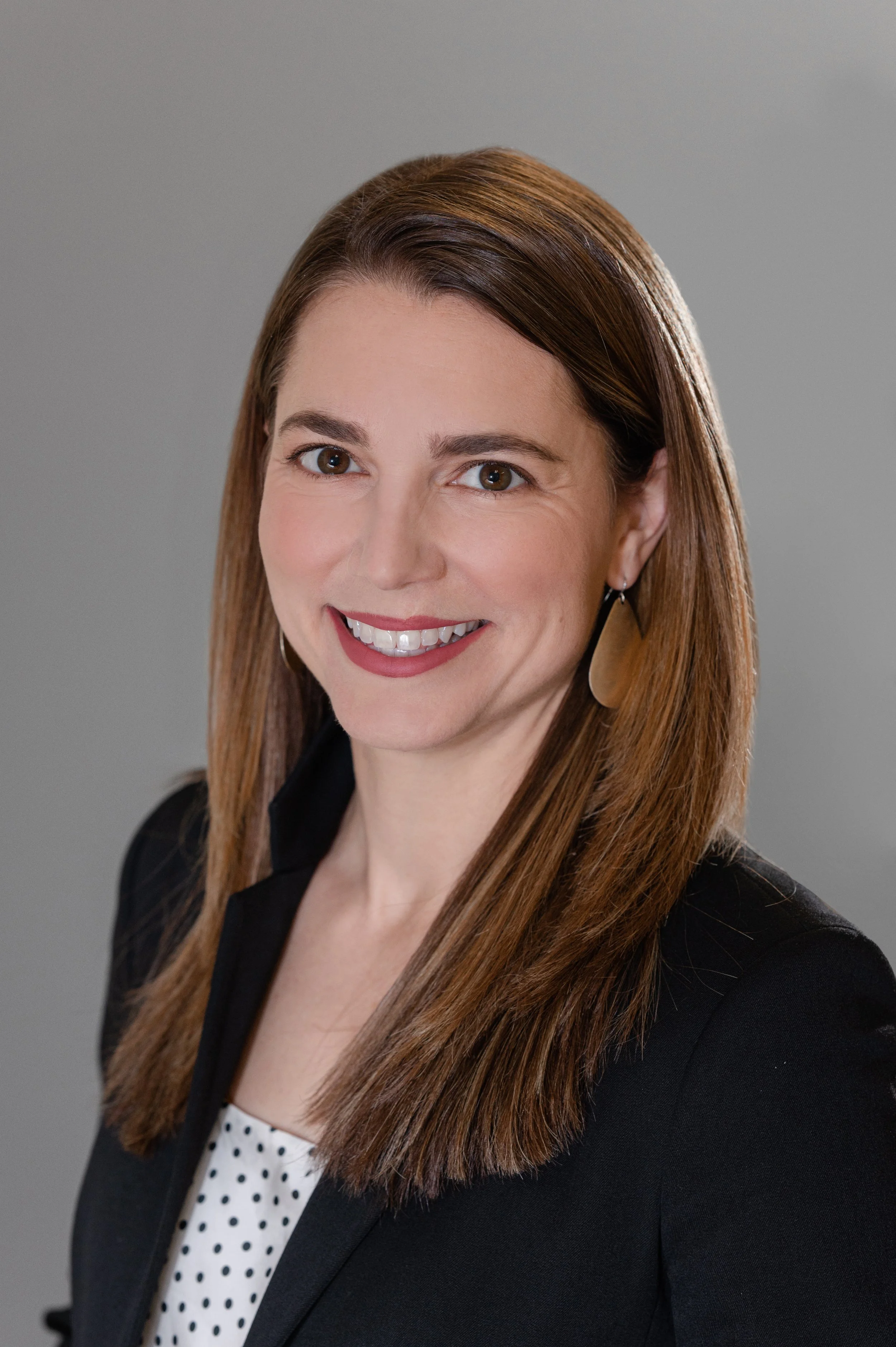 A woman with long brown hair smiling, wearing a black blazer over a white and black polka dot top, and a tan earring, against a plain gray background.