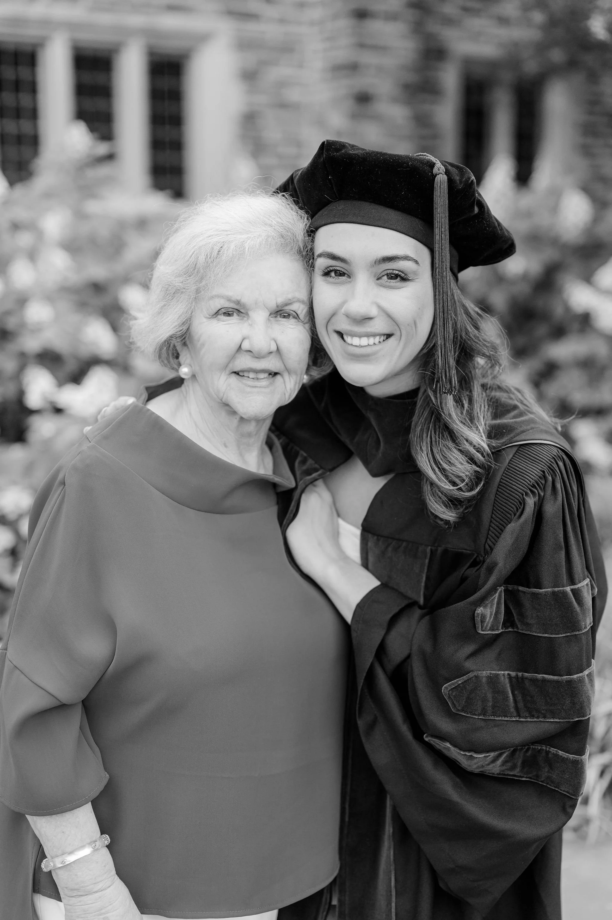 A young woman in a graduation gown and cap, smiling, with an elderly woman, both embracing outdoors.