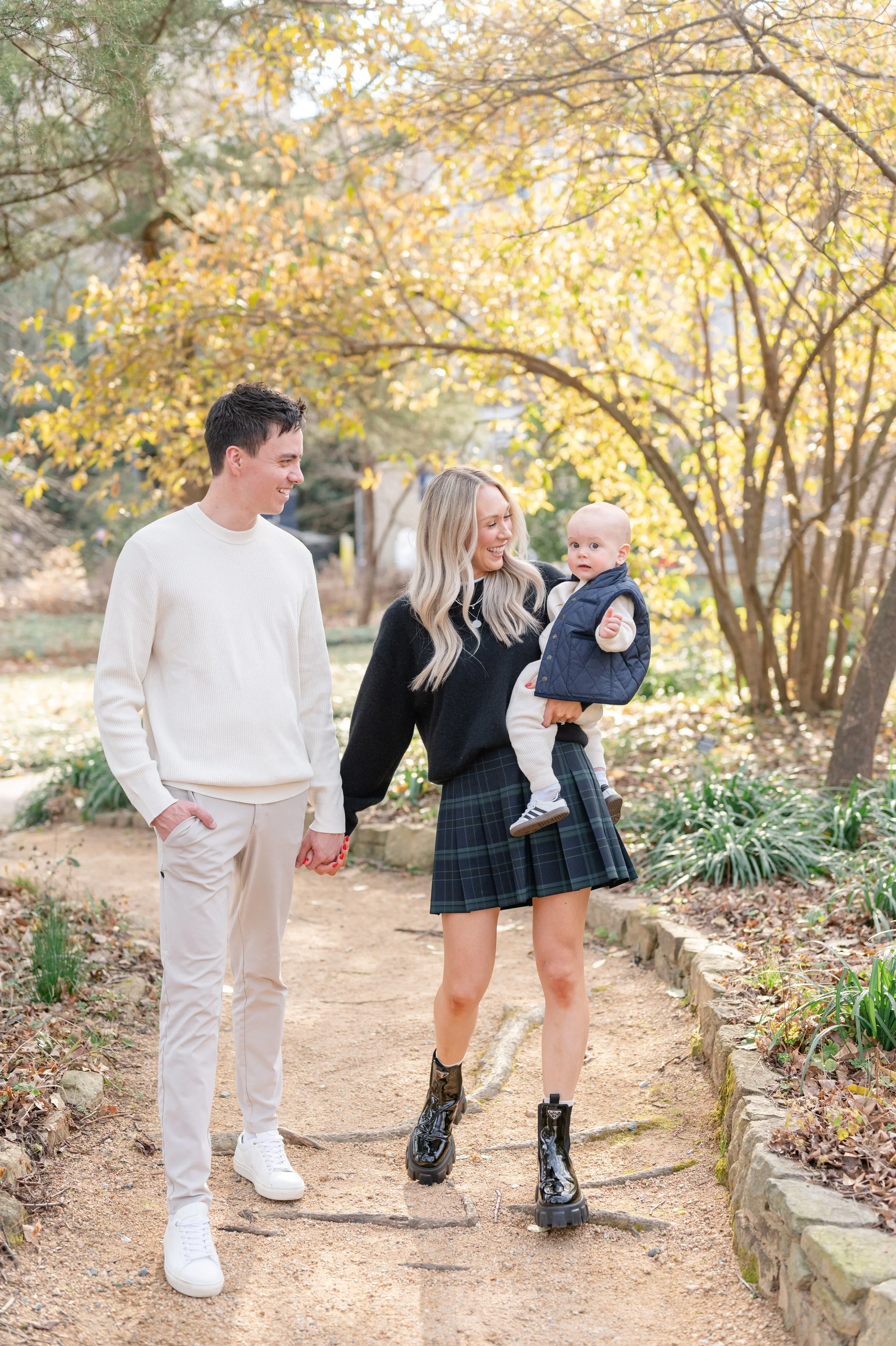 A woman holding a baby and a man holding hands walking in a park with trees and fall foliage.