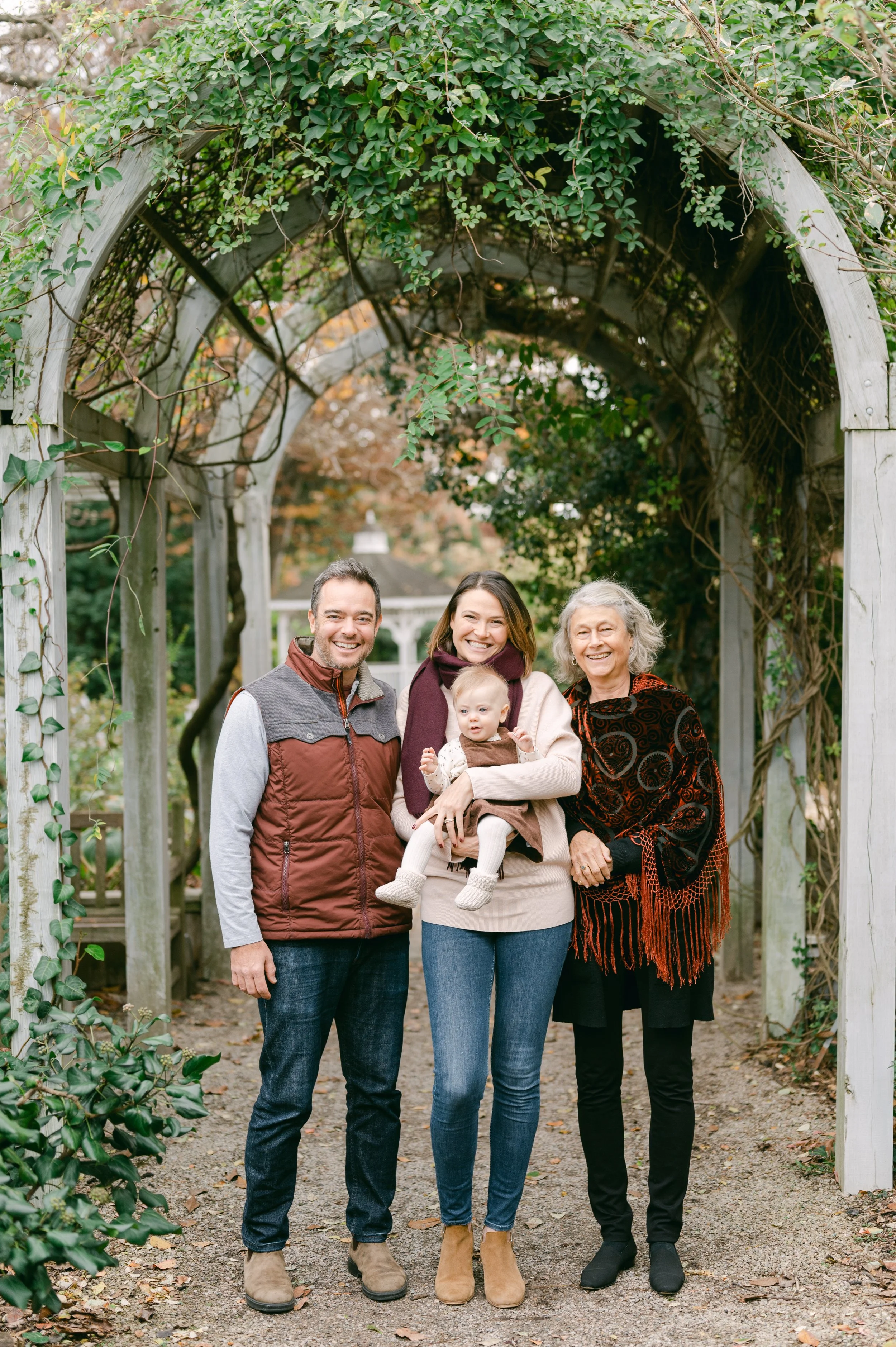 A multi-generational family of five standing under a wooden arbor with greenery, smiling, dressed in casual fall clothing, in a garden or park setting.