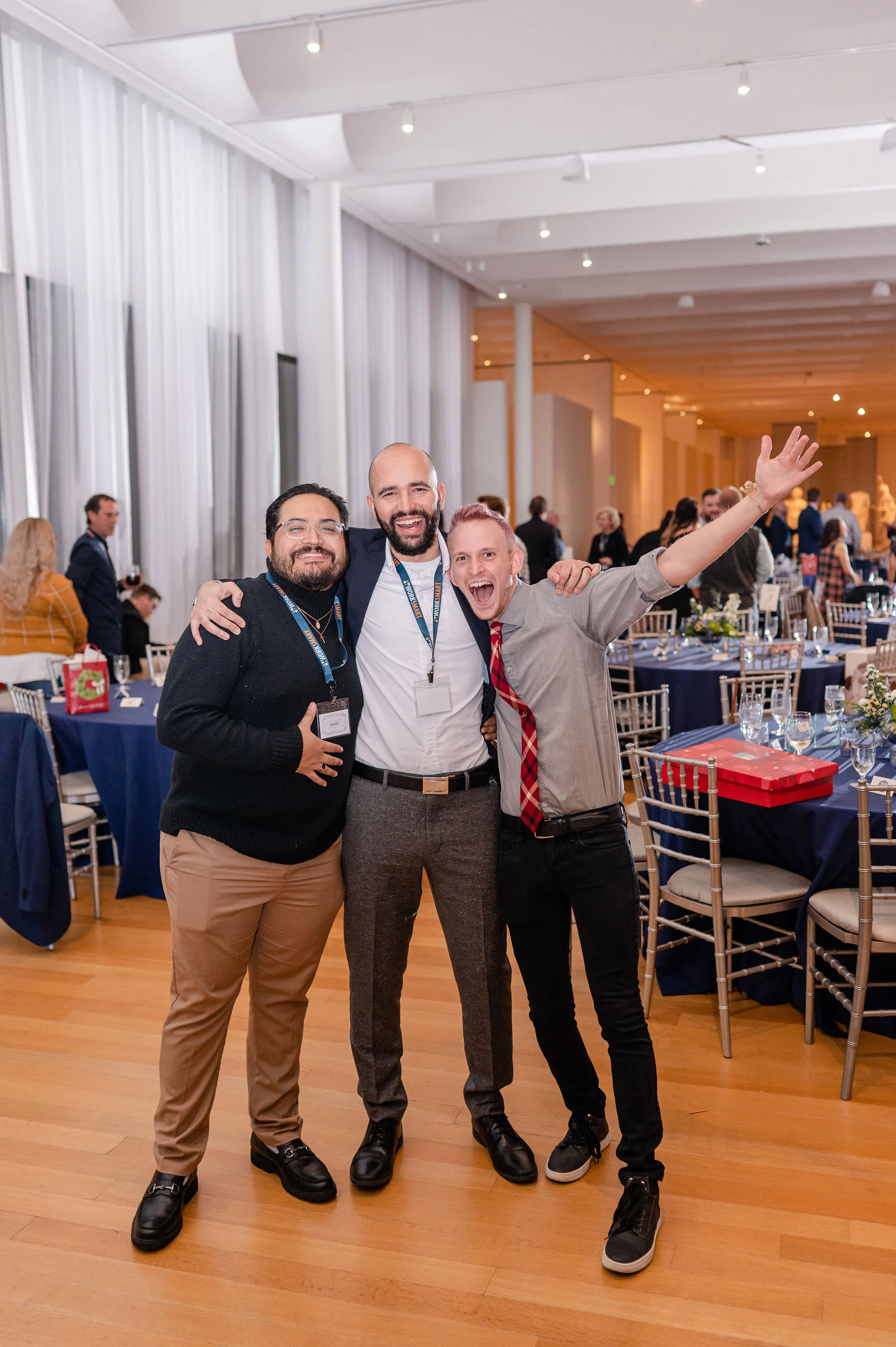 Three men standing together indoors at a social event, smiling and posing for the camera. The man in the middle has his arms around the other two, one making a peace sign and the other holding a drink.