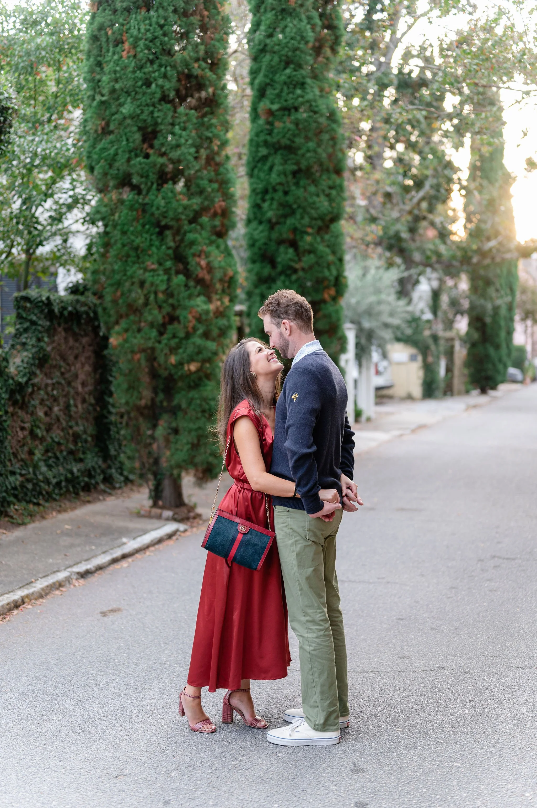 A romantic couple standing close together on a quiet street, with trees and houses in the background during sunset, sharing an intimate moment and smiling at each other.