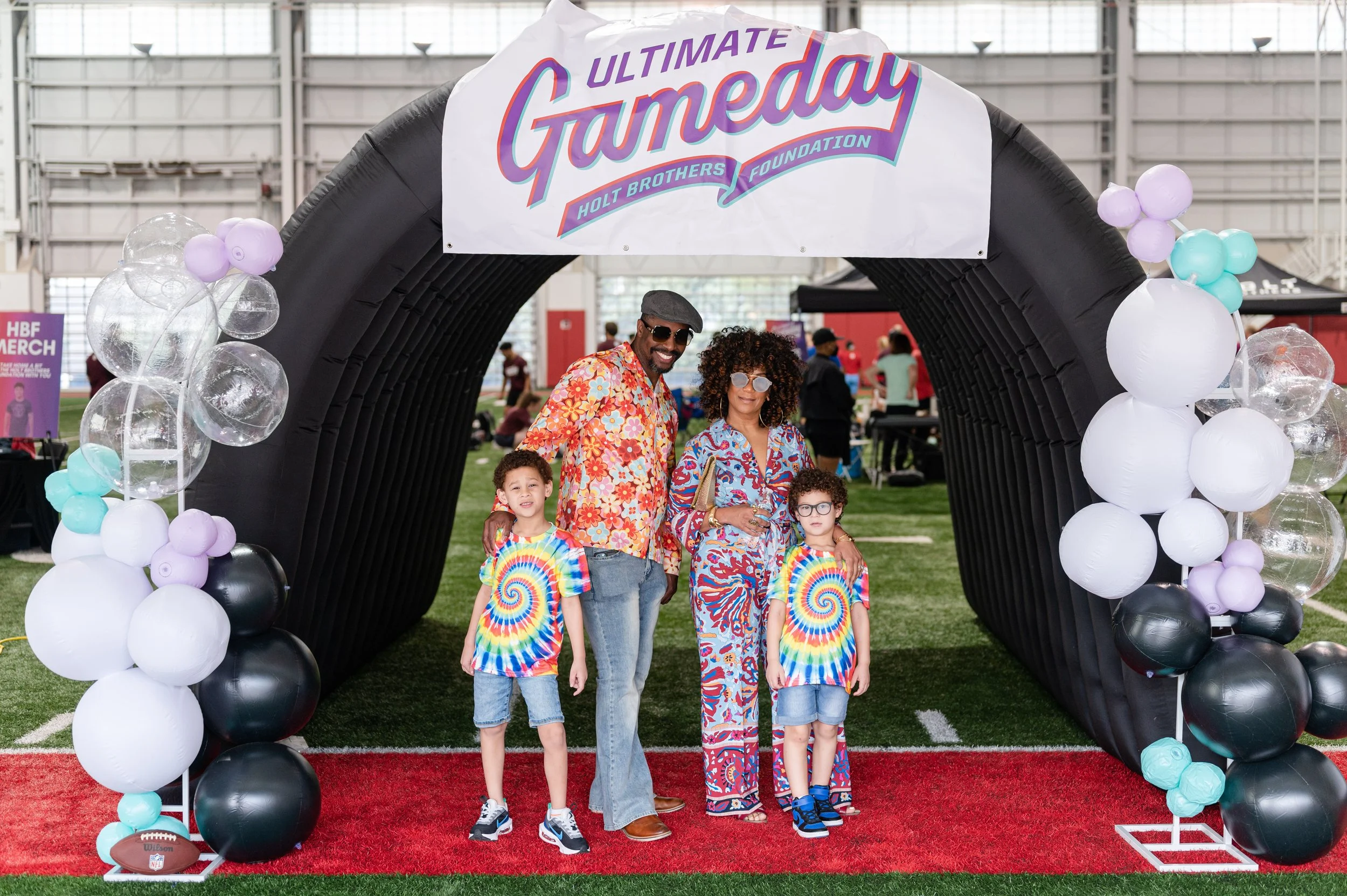 Family of four at a themed event under a black inflatable arch decorated with pastel balloons, with a sign saying 'Ultimate Game Day' and 'Holt Brothers Foundation' in a large indoor venue.