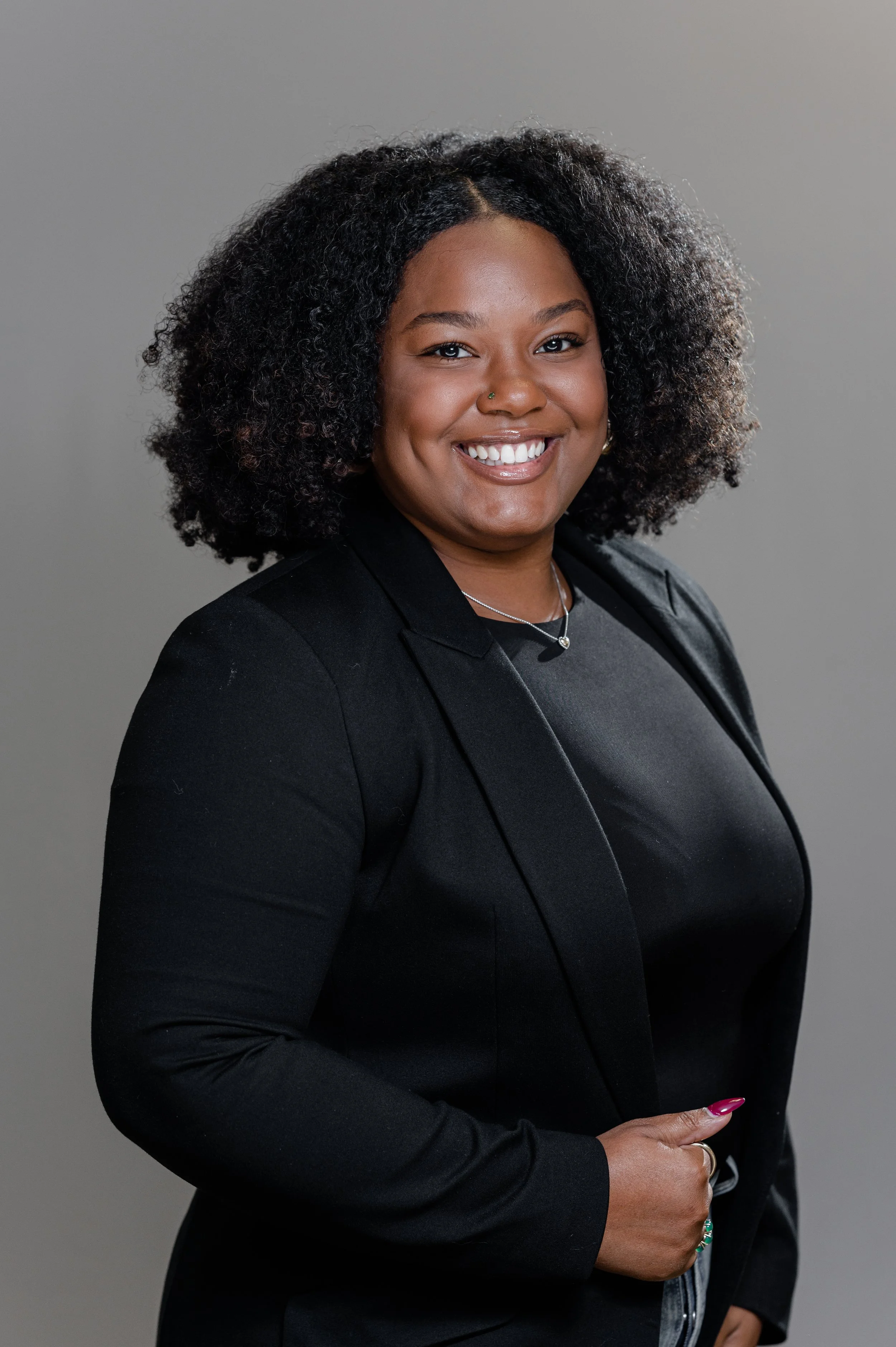 A woman with curly hair, smiling, wearing a black blazer and top, standing against a gray background.