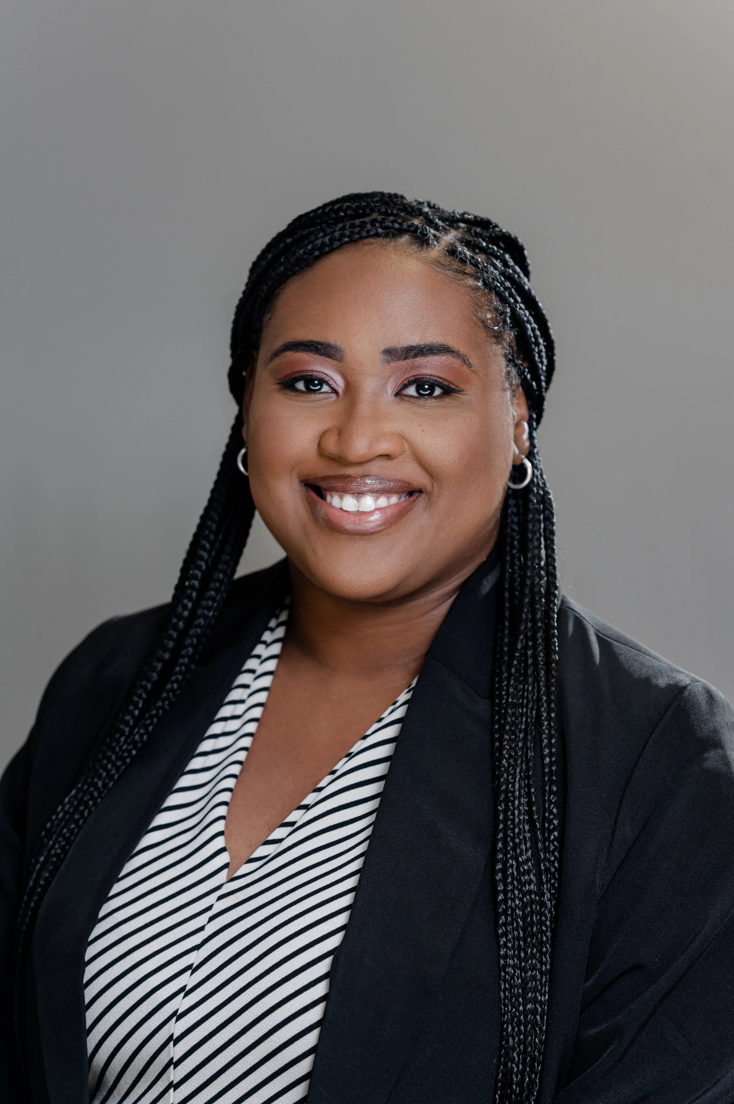 Professional portrait of a black woman with braided hair, smiling, wearing hoop earrings, a black blazer over a black and white striped shirt, against a plain gray background.