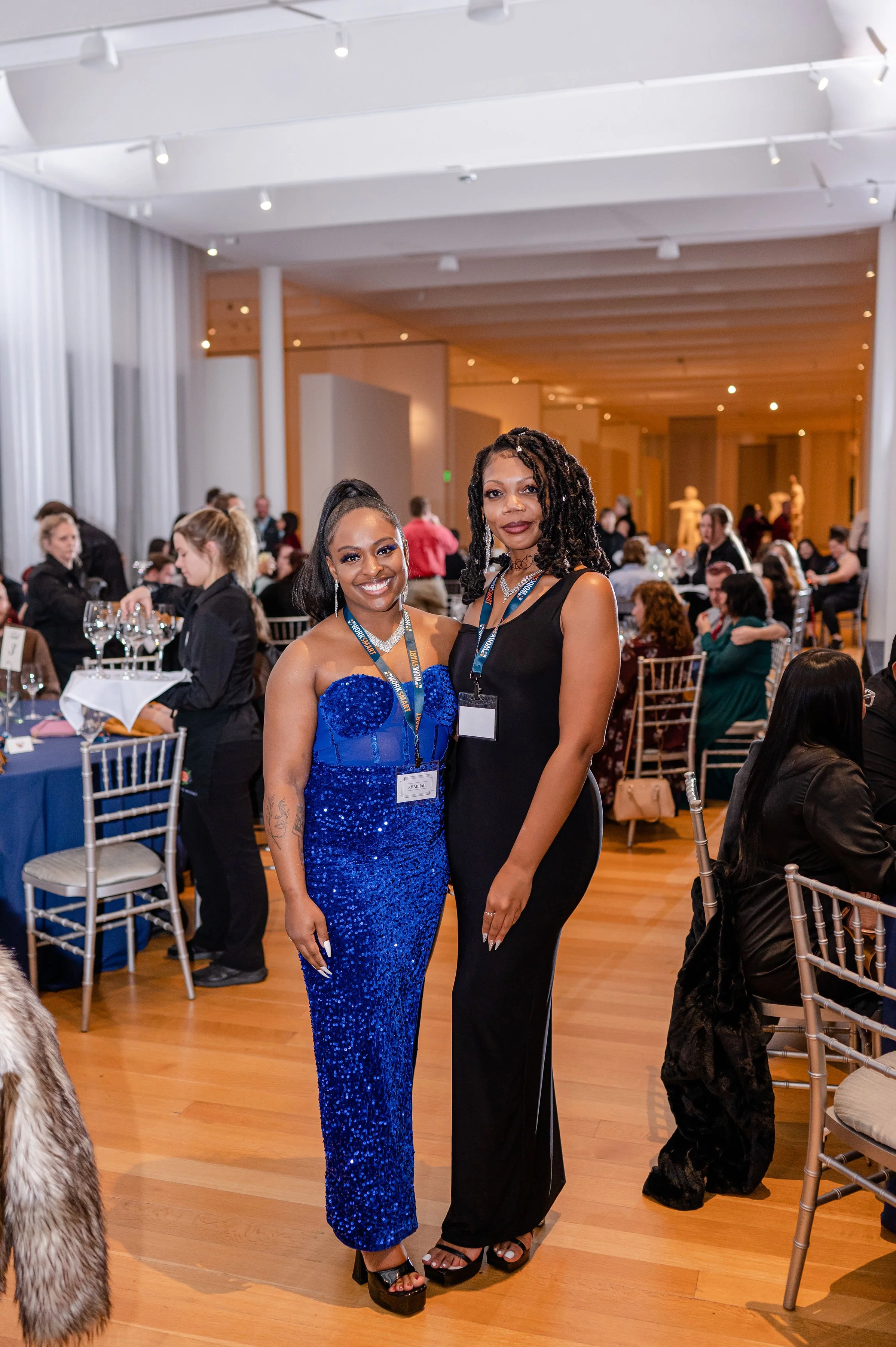 Two black women dressed in elegant evening wear standing together at a banquet or conference in a large, well-lit room with many seated attendees and decorated tables.