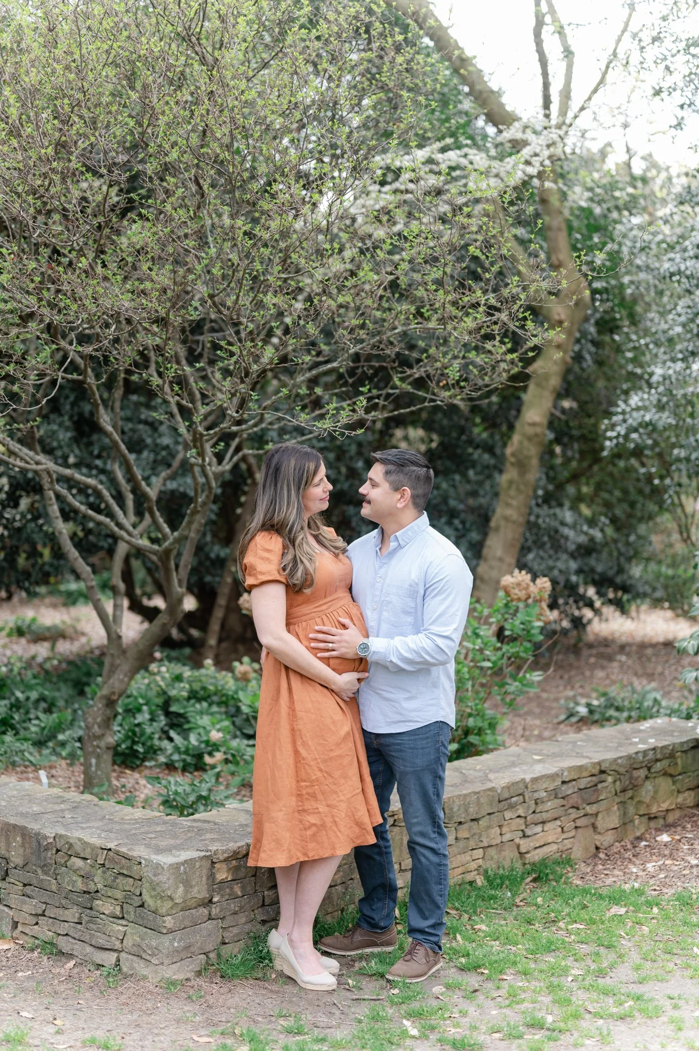 A pregnant woman in an orange dress and a man in a light blue shirt stand closely, looking into each other's eyes affectionately, while standing outside on a stone pathway surrounded by greenery and blooming trees.