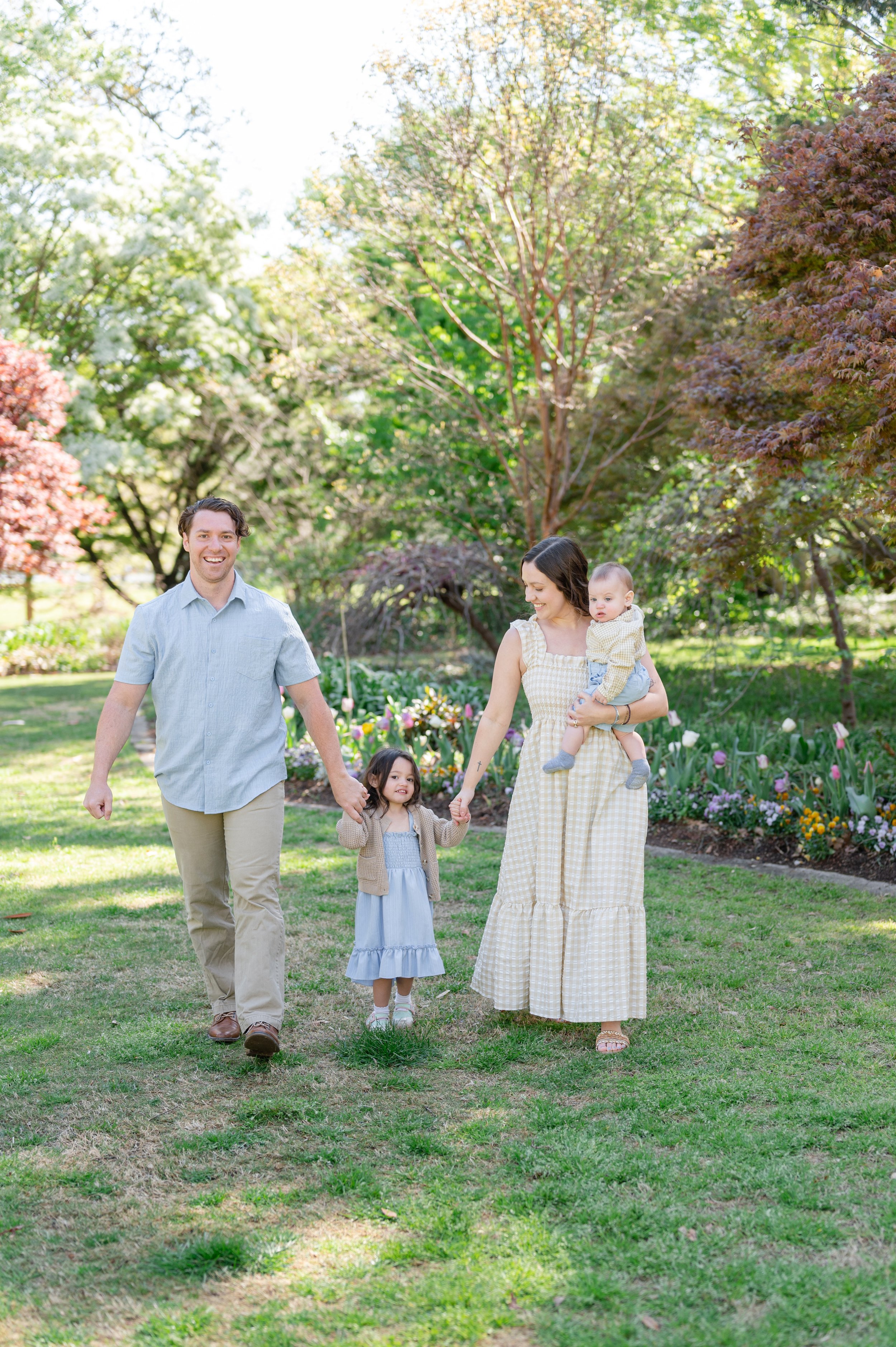 A family of four walking in a garden with colorful flowers and trees. The father has brown hair and wears a light blue shirt and beige pants. The mother has dark hair and wears a yellow checkered dress, carrying a baby in her arms. The baby has light hair and is dressed in yellow. Their daughter has dark hair, wears a light blue dress, and holds hands with her father and mother.
