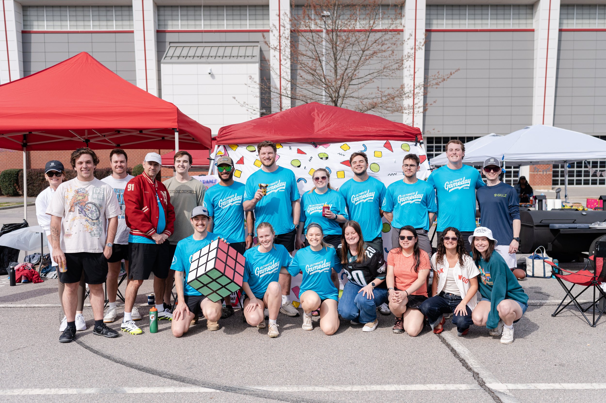 A group of diverse young people smiling and posing together outdoors at a community event. They are standing in front of tents and a building, some holding drinks and a large Rubik's cube.