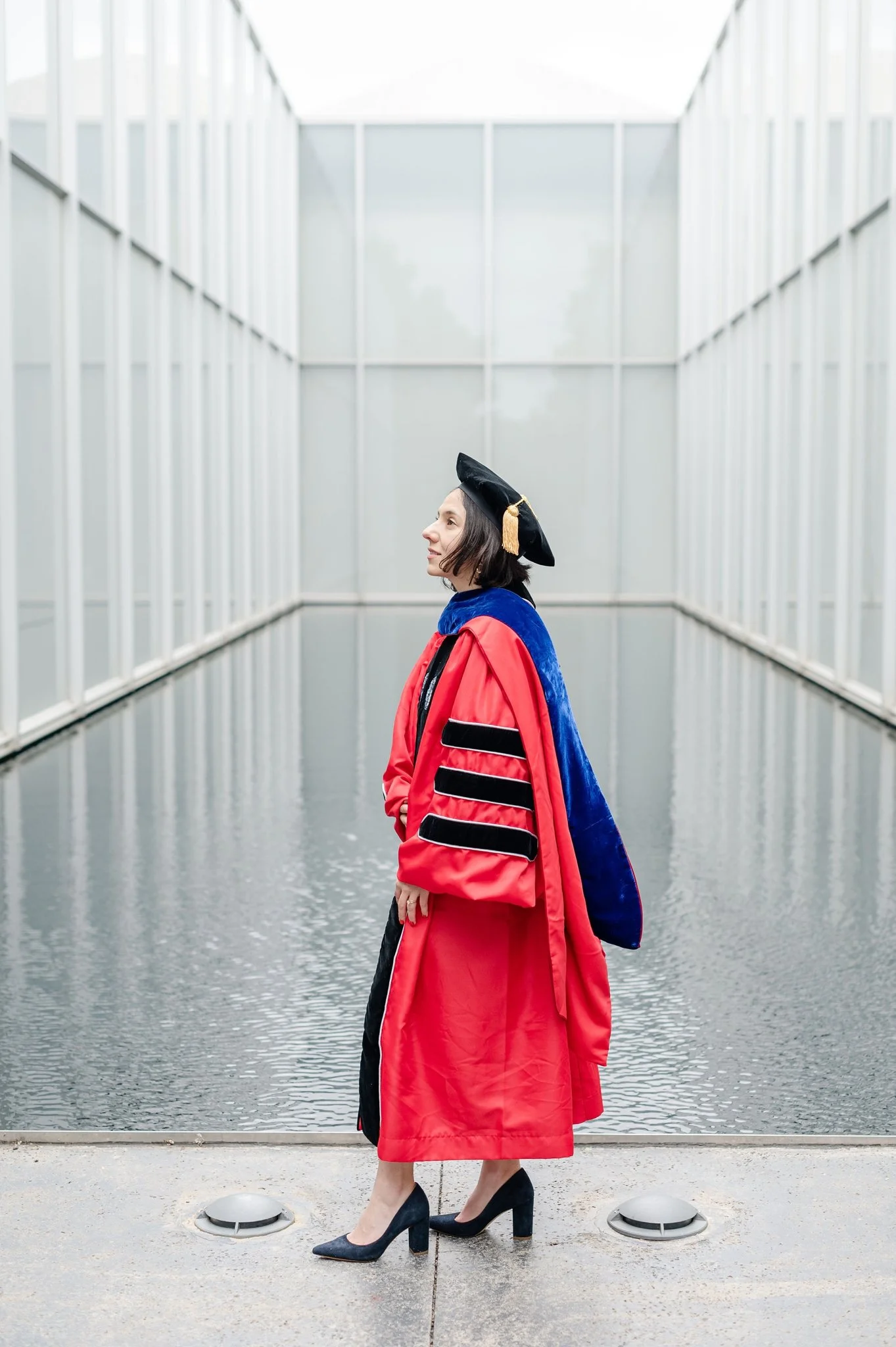 A woman in a black academic cap with a tassel, wearing a red gown with black accents, stands beside water inside a modern, glass-sided corridor.