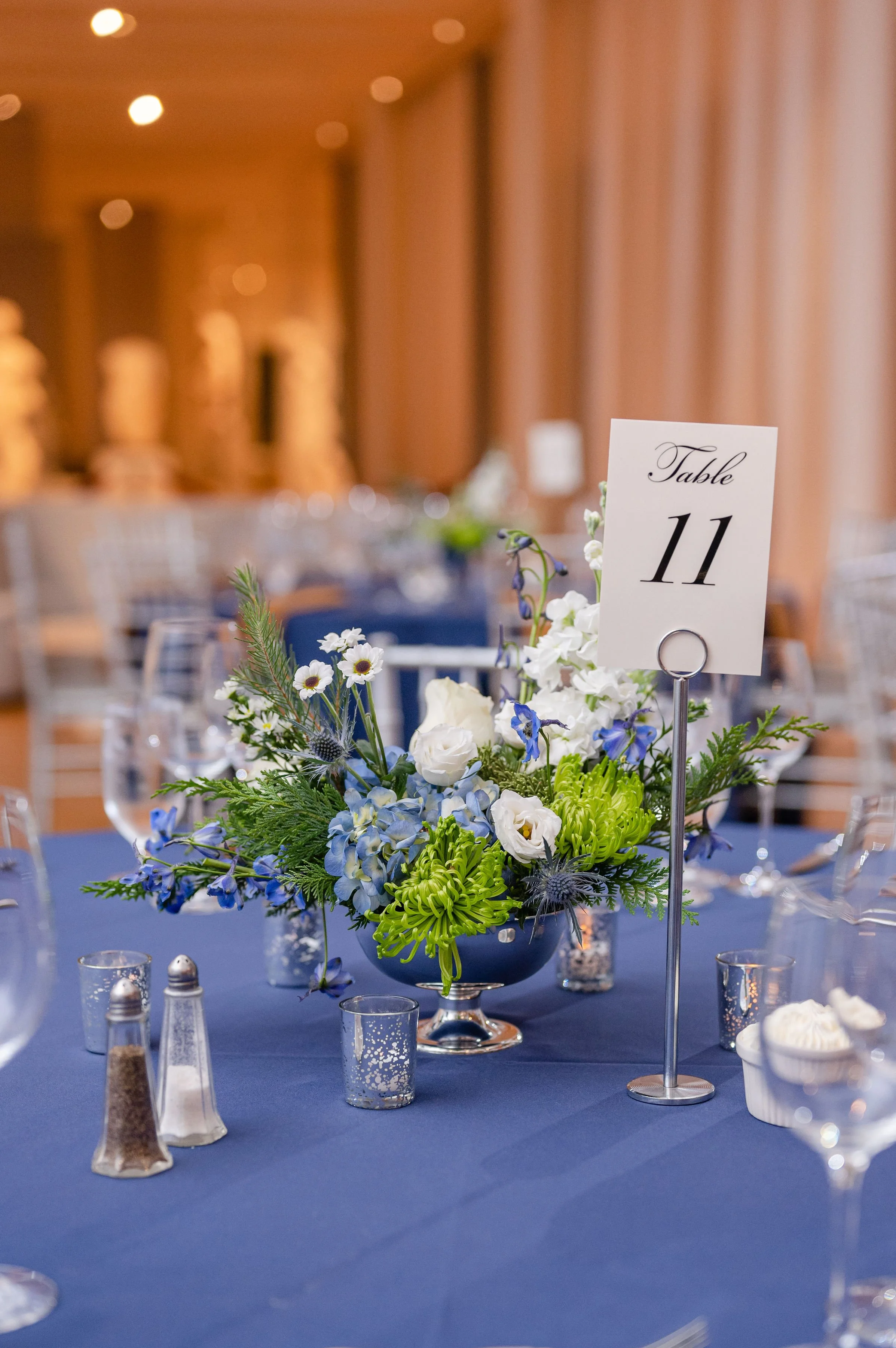 A floral centerpiece with white, blue, and green flowers on a blue tablecloth at a formal event, with a small table number sign indicating 'Table 11'.