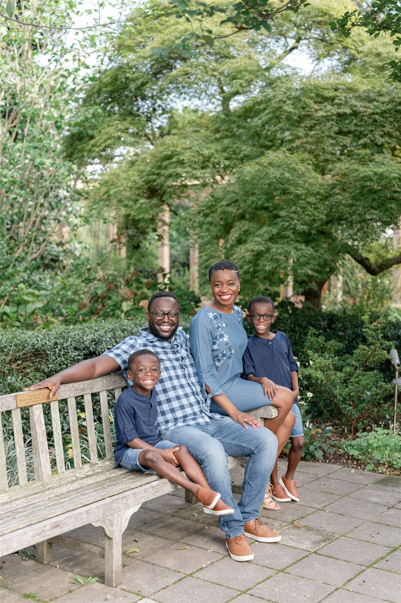 A happy black family of four, including two adults and two children, sitting on a wooden park bench surrounded by lush green trees and bushes during daytime.