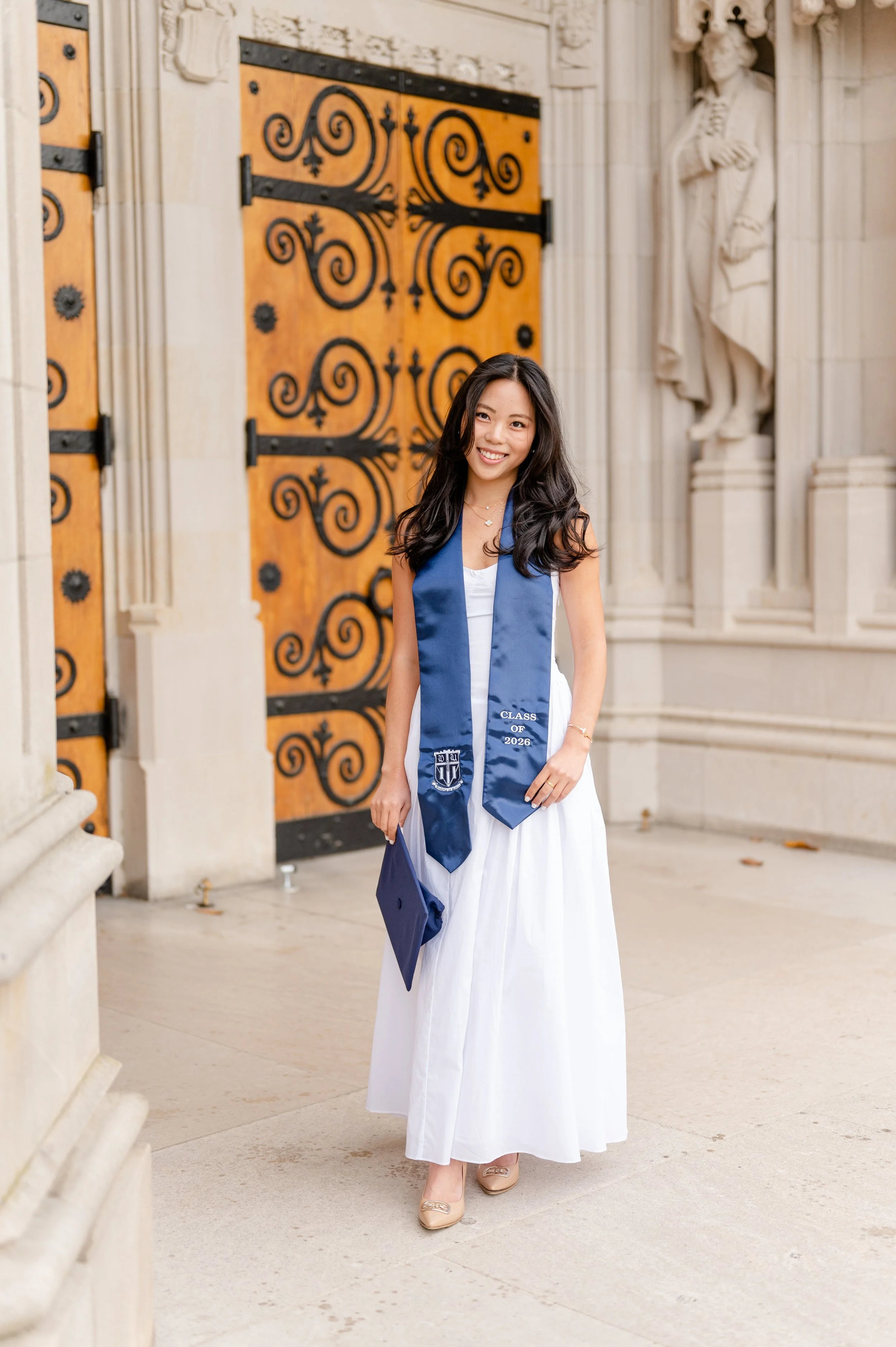 Young woman in a white dress and beige heels, graduation cap in hand, standing outside a building with ornate wooden door and stone columns.