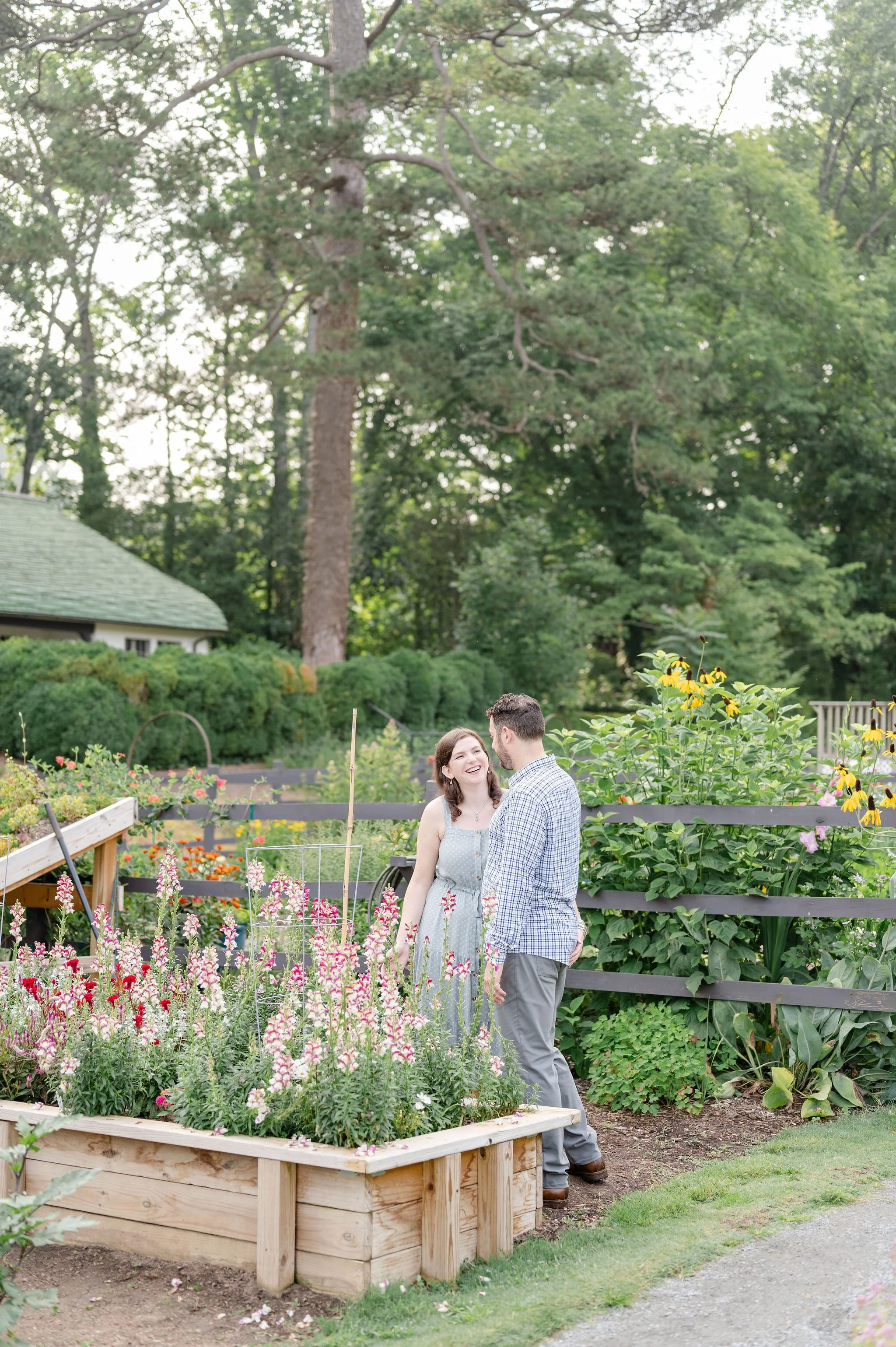 Couple standing in a lush garden filled with colorful flowers, surrounded by green trees and plants, with a wooden fence in the background.