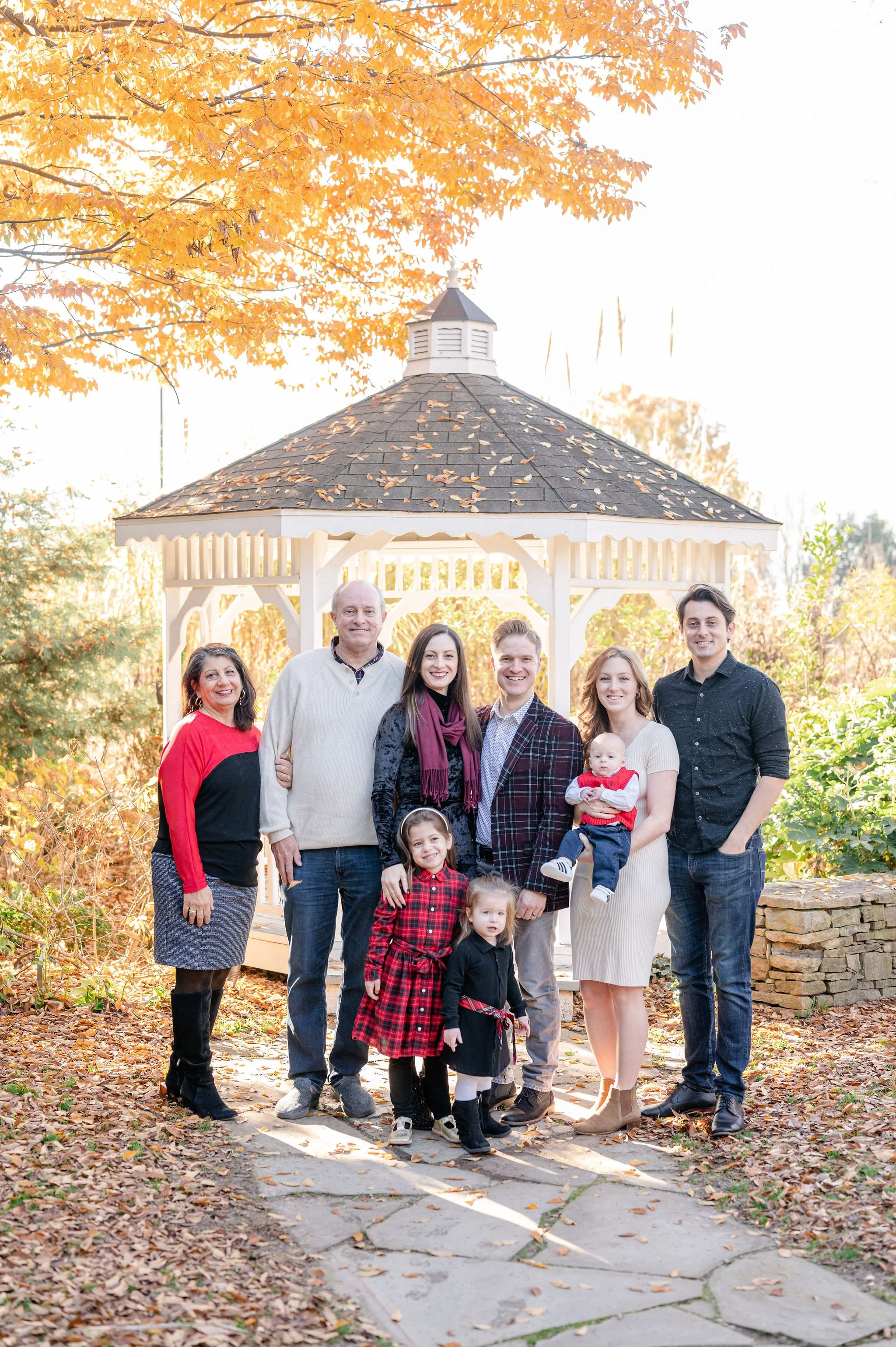 A multi-generational family gathering outdoors in autumn, standing on a stone pathway with fallen leaves, under a gazebo, surrounded by trees with orange and yellow leaves, smiling at the camera.