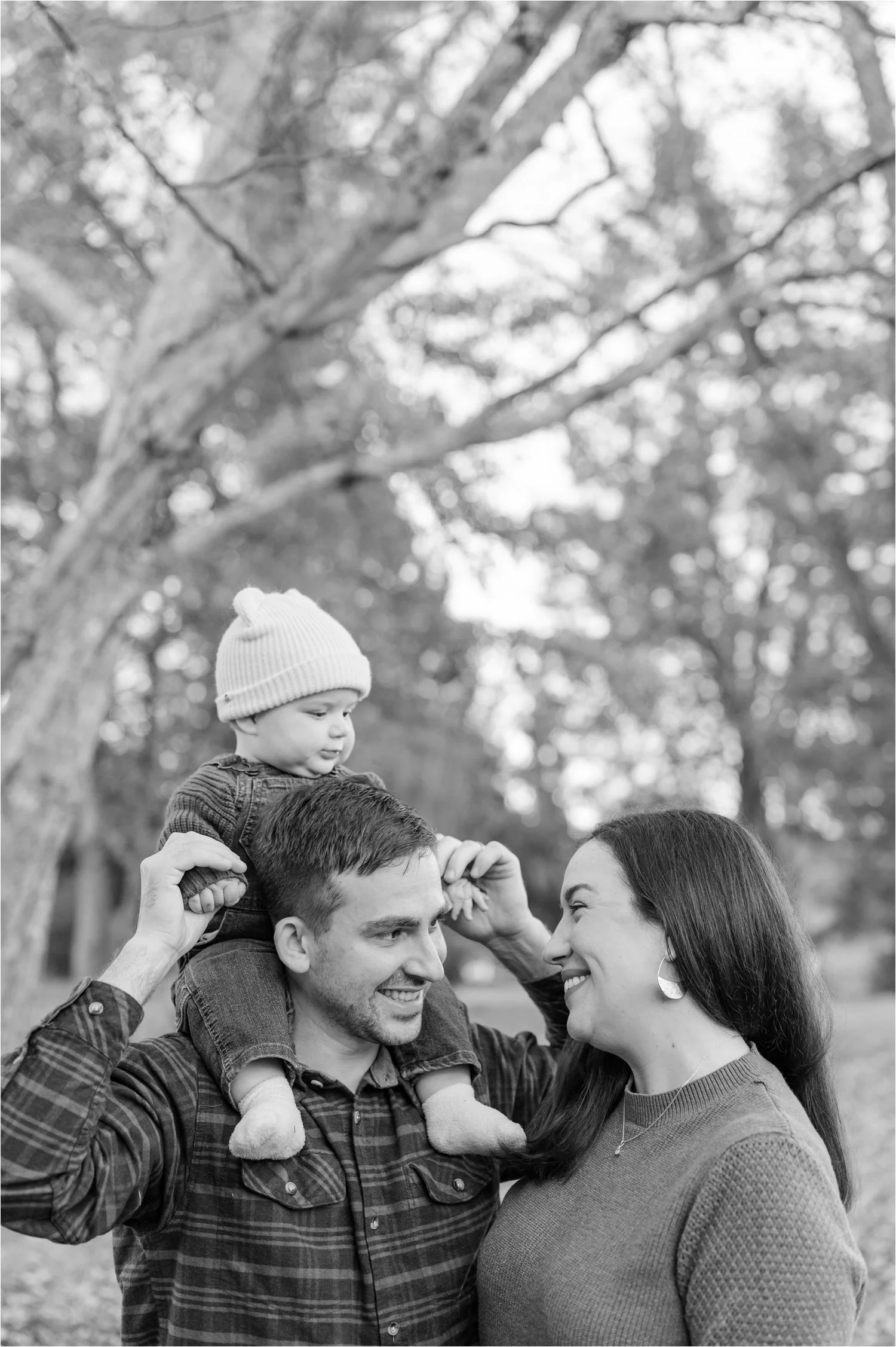 A man with a beard and a woman with long dark hair smiling at each other as they hold a young boy on his shoulders in a park with trees in the background.