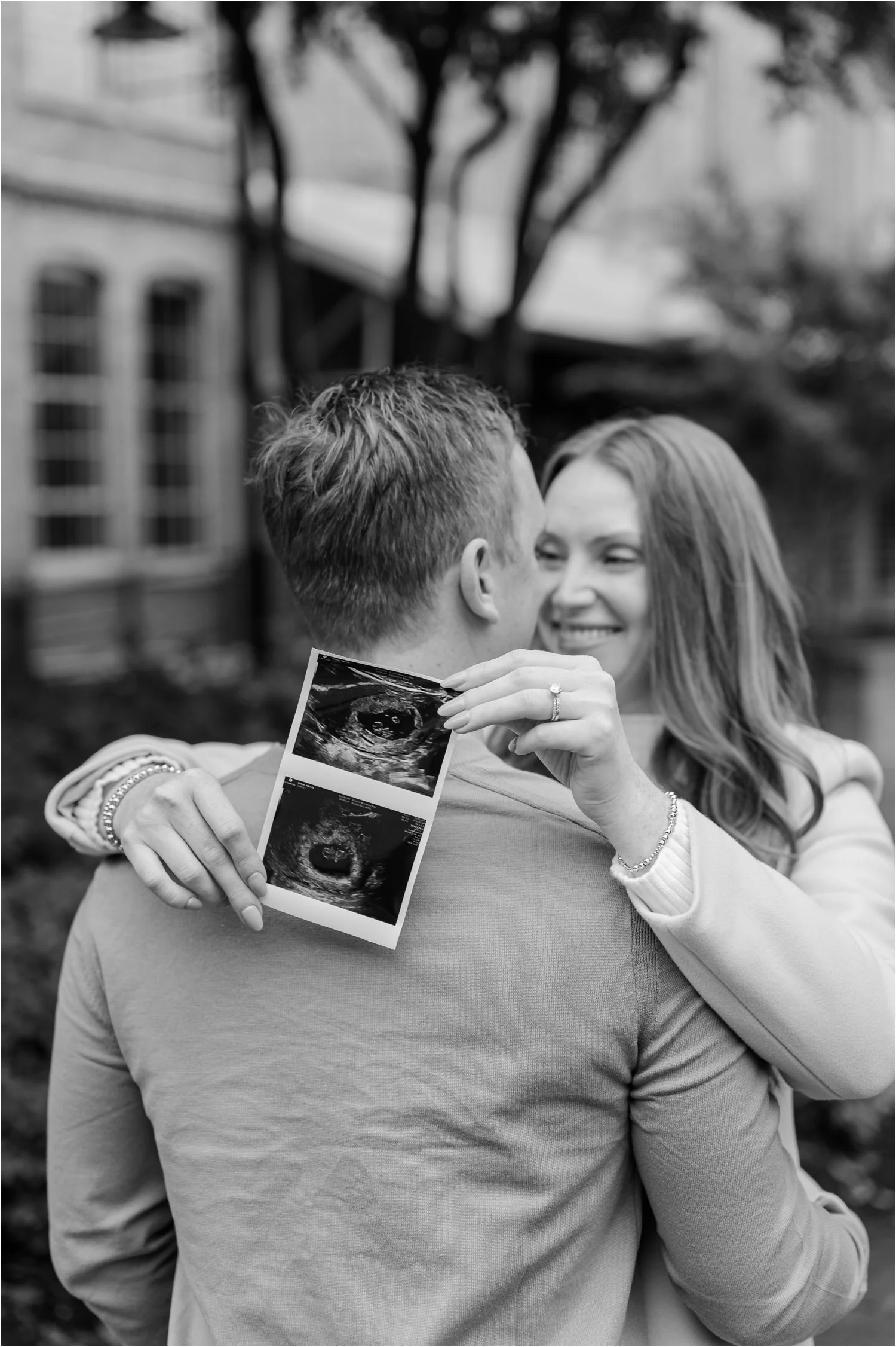 A couple sharing a kiss outdoors. The woman is holding an ultrasound picture, smiling as the man embraces her.