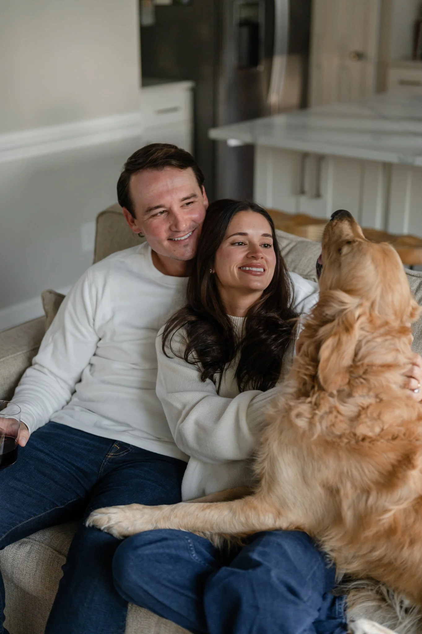 A happy couple sitting on a couch in their living room, smiling and cuddling with their large golden retriever dog.