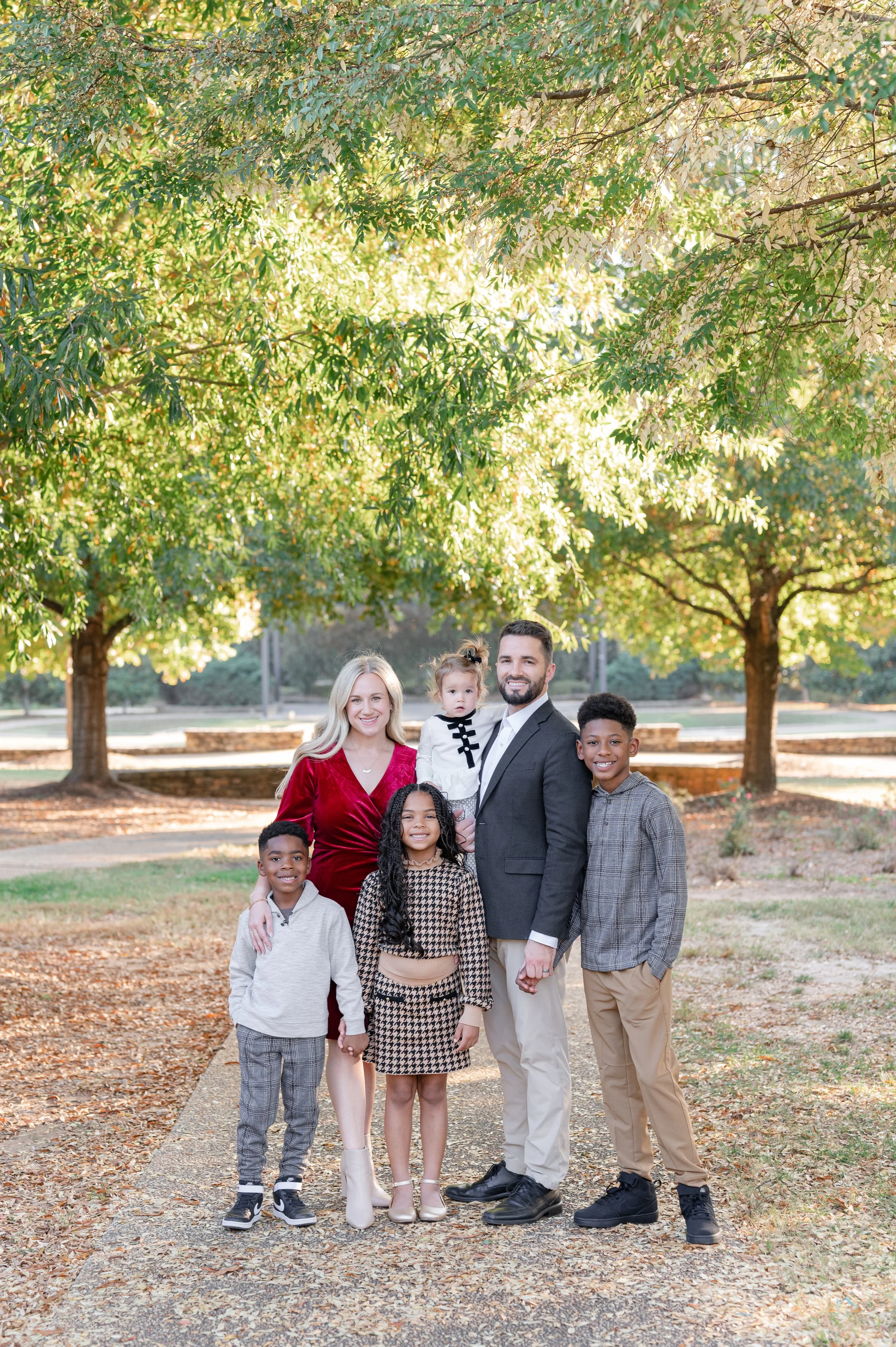 A diverse family group of six people, including black children and adults, standing outdoors on a walkway in a park with green trees and sunlight filtering through the leaves.