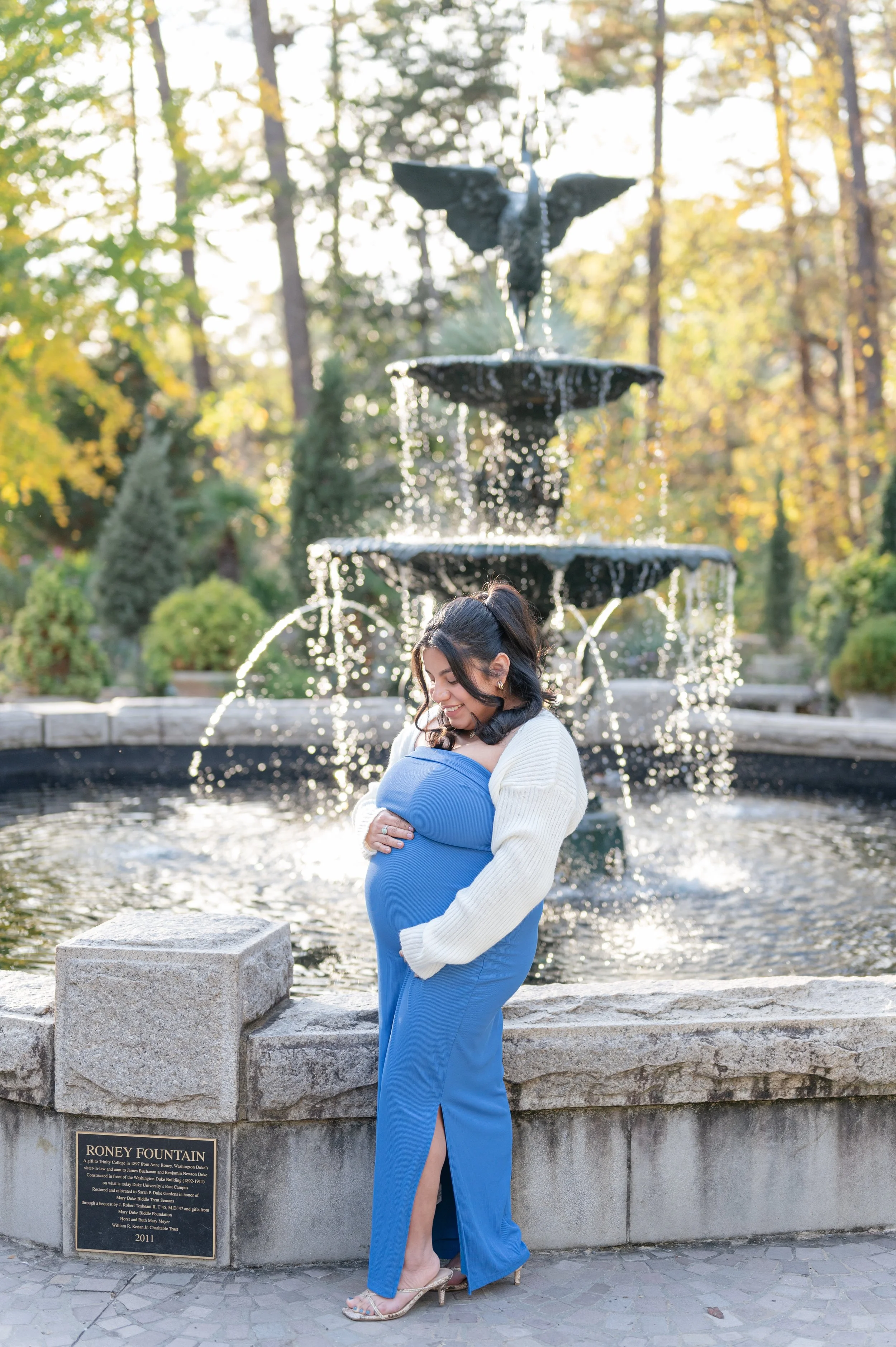 A pregnant woman in a blue dress and white cardigan standing in front of a fountain and smiling while cradling her belly in Roney Fountain, a park or garden setting with trees and greenery