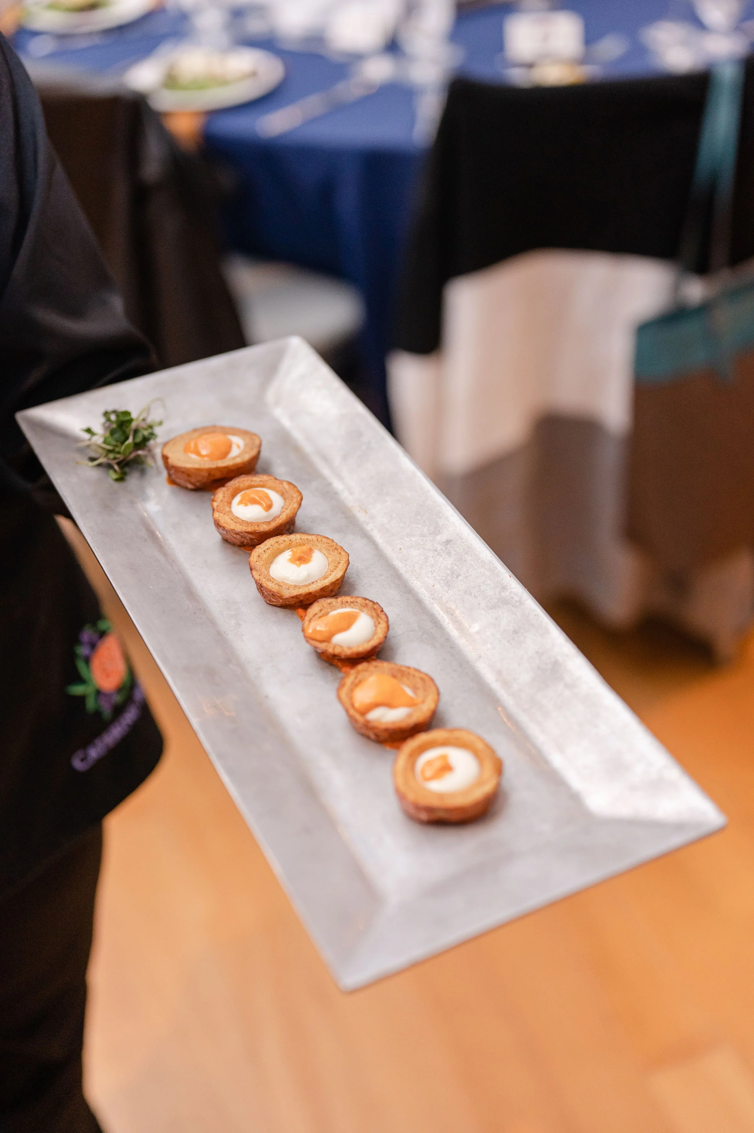 A person holding a rectangular plate with a row of small, round appetizers topped with a dollop of cream and orange sauce, garnished with microgreens, set against a background of a table with more plates at a formal event.