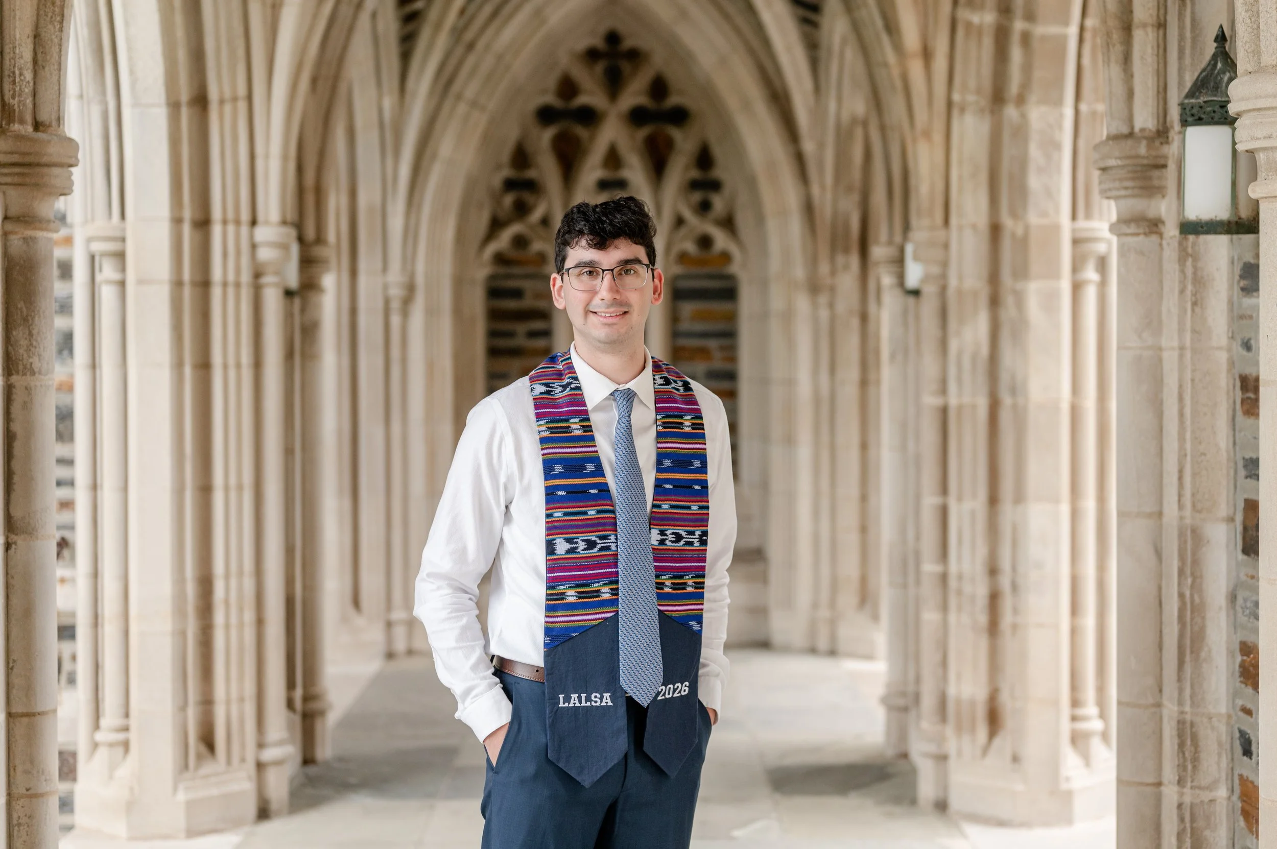 Young man in a white shirt with a patterned stole, glasses, and dark pants stands in a stone archway, smiling, during a graduation ceremony.