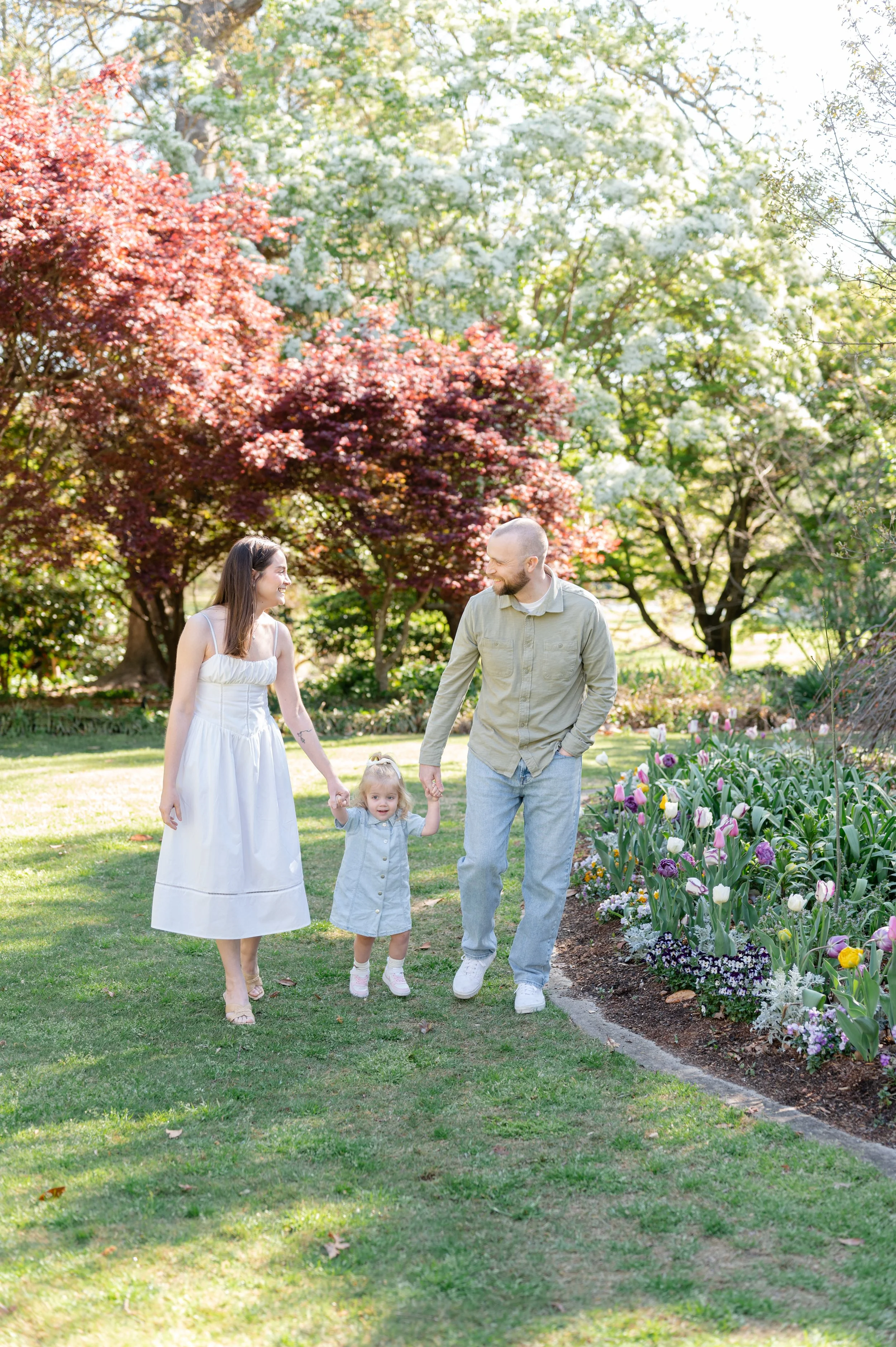 A family of three walking through a garden with blooming flowers, trees, and bright sunlight.