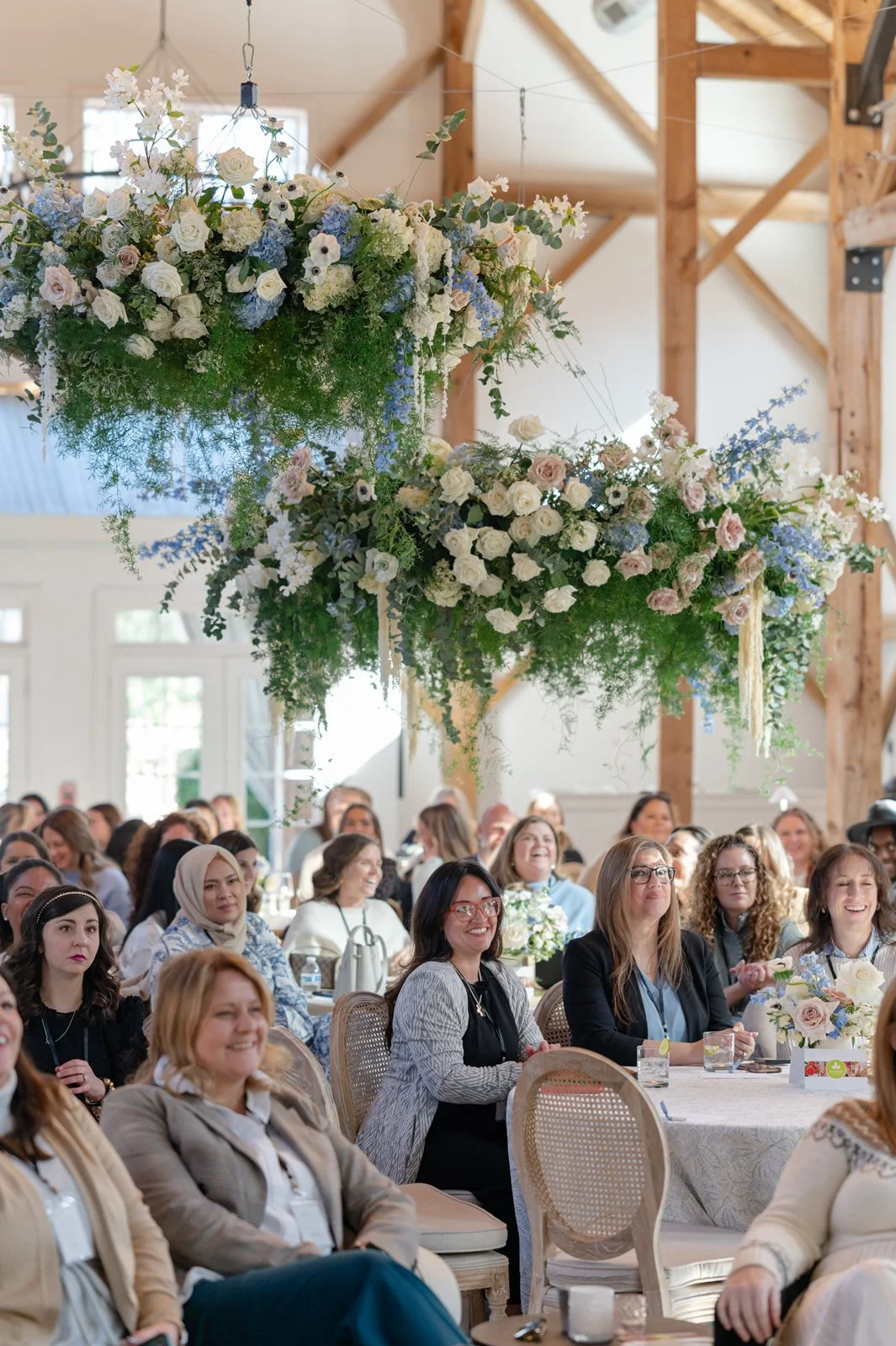A large floral arrangement with white, blue, and light pink flowers hanging above a diverse group of women seated at tables during a conference or event in a spacious, well-lit room with exposed wood beams.