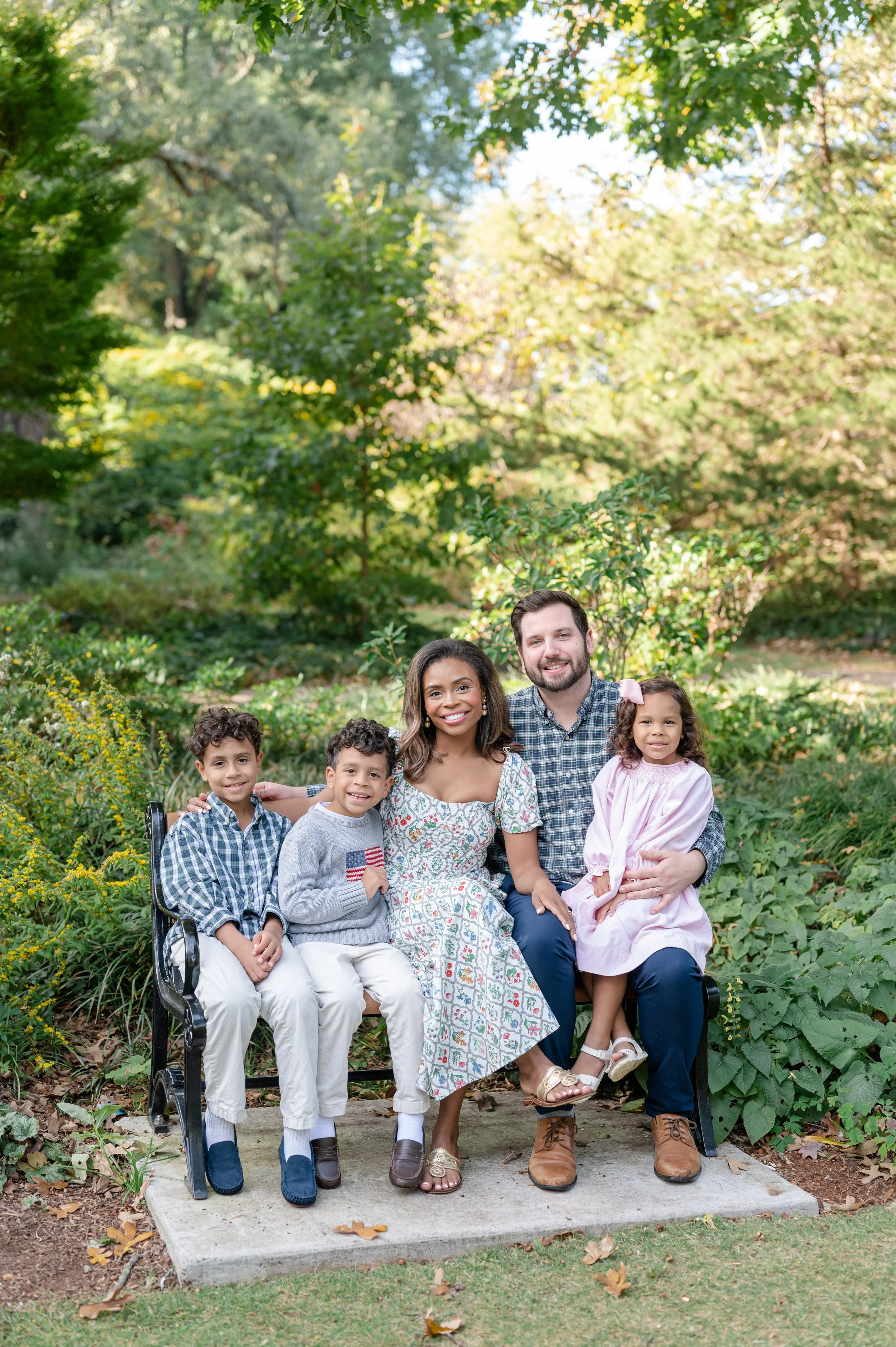 A family of five sitting on a bench outdoors among greenery, smiling at the camera.