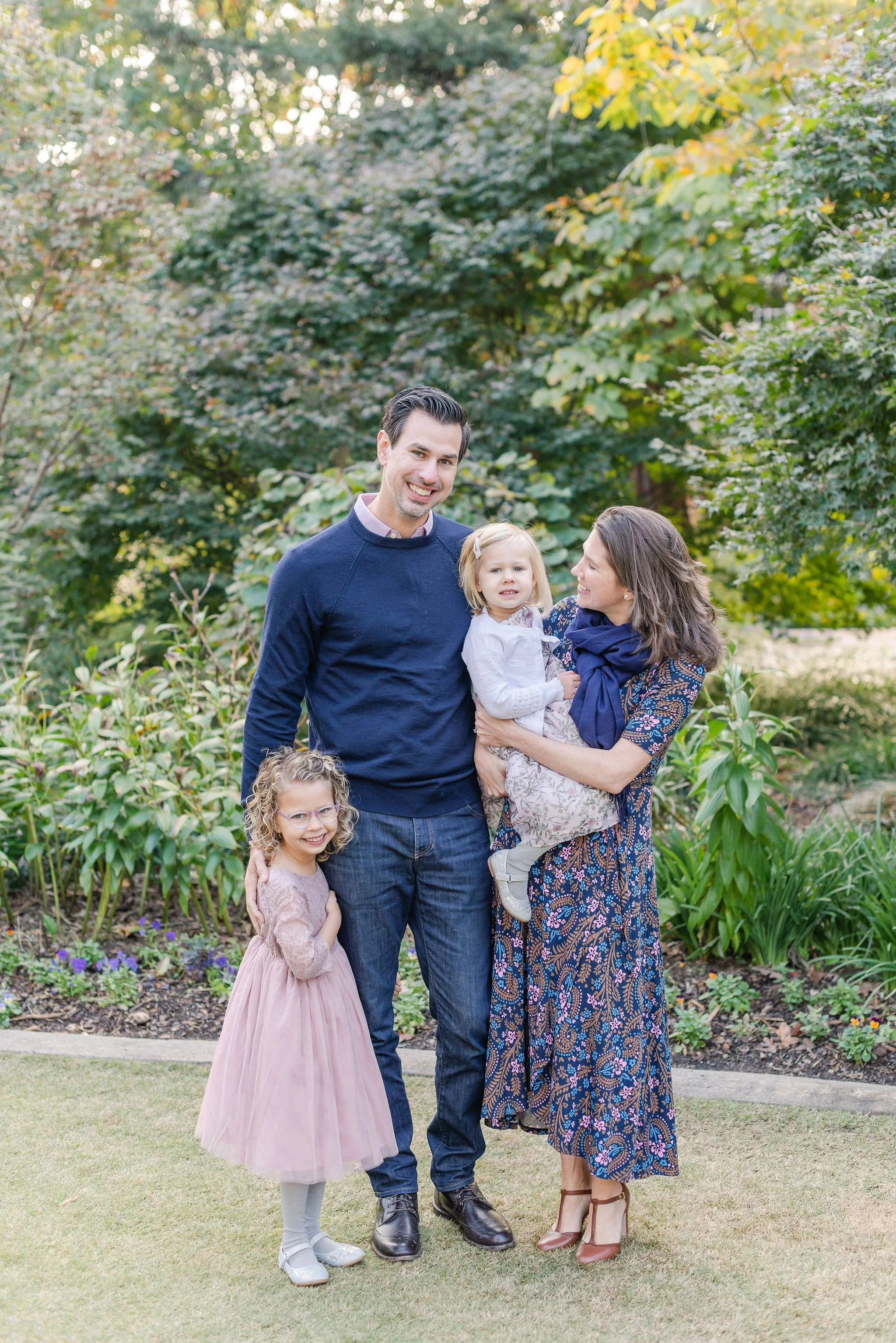 A family of five standing outdoors in a garden with green trees and plants. The man is smiling and wearing a navy blue sweater and jeans. The woman is holding a young girl, both are smiling; she is wearing a floral dress and the girl a white top with a patterned skirt. A young girl with curly hair and glasses is standing in front, wearing a pink dress and white tights. There is another child with curly hair at the bottom left corner, partially visible, wearing glasses and a light-colored dress.