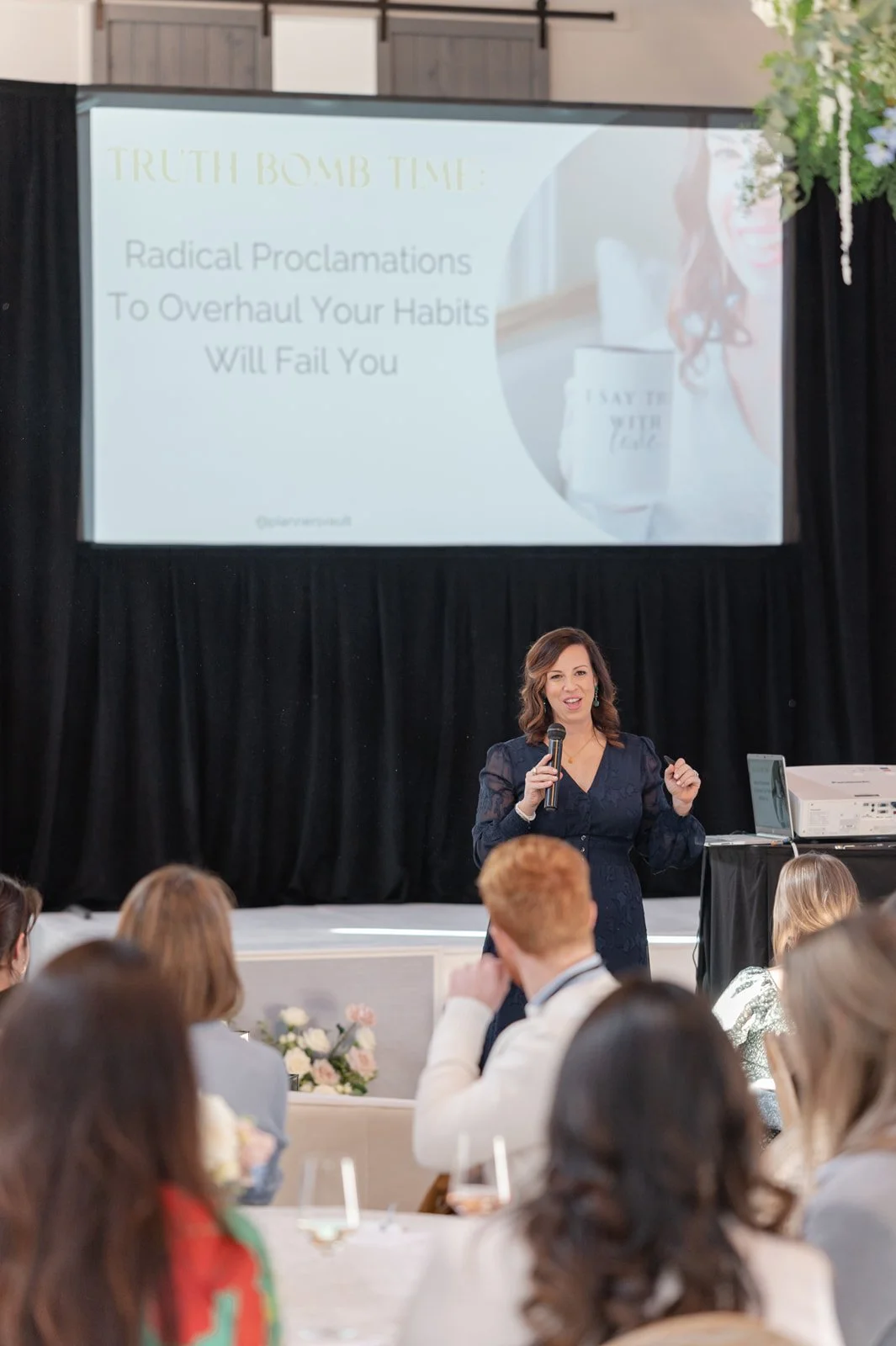 A woman in a dark dress speaking into a microphone during a presentation at a conference, with a large screen behind her displaying a slide titled 'TRUTH BOMB TIME' with the subtitle 'Radical Proclamations To Overhaul Your Habits Will Fail You'. Several audience members are seated at tables, listening attentively.