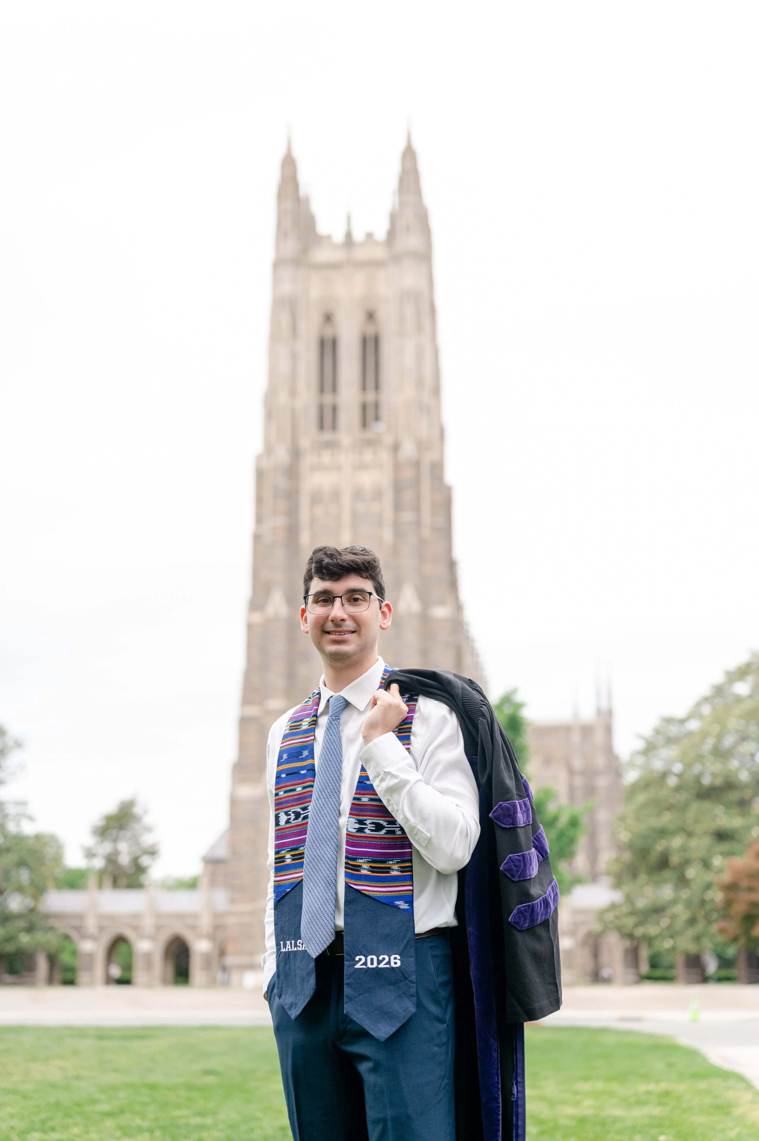 A young man in a white dress shirt, blue tie, and graduation robes standing outdoors on a university campus with a historic building in the background. He carries a backpack over his shoulder and wears a colorful stole around his neck, smiling at the camera.