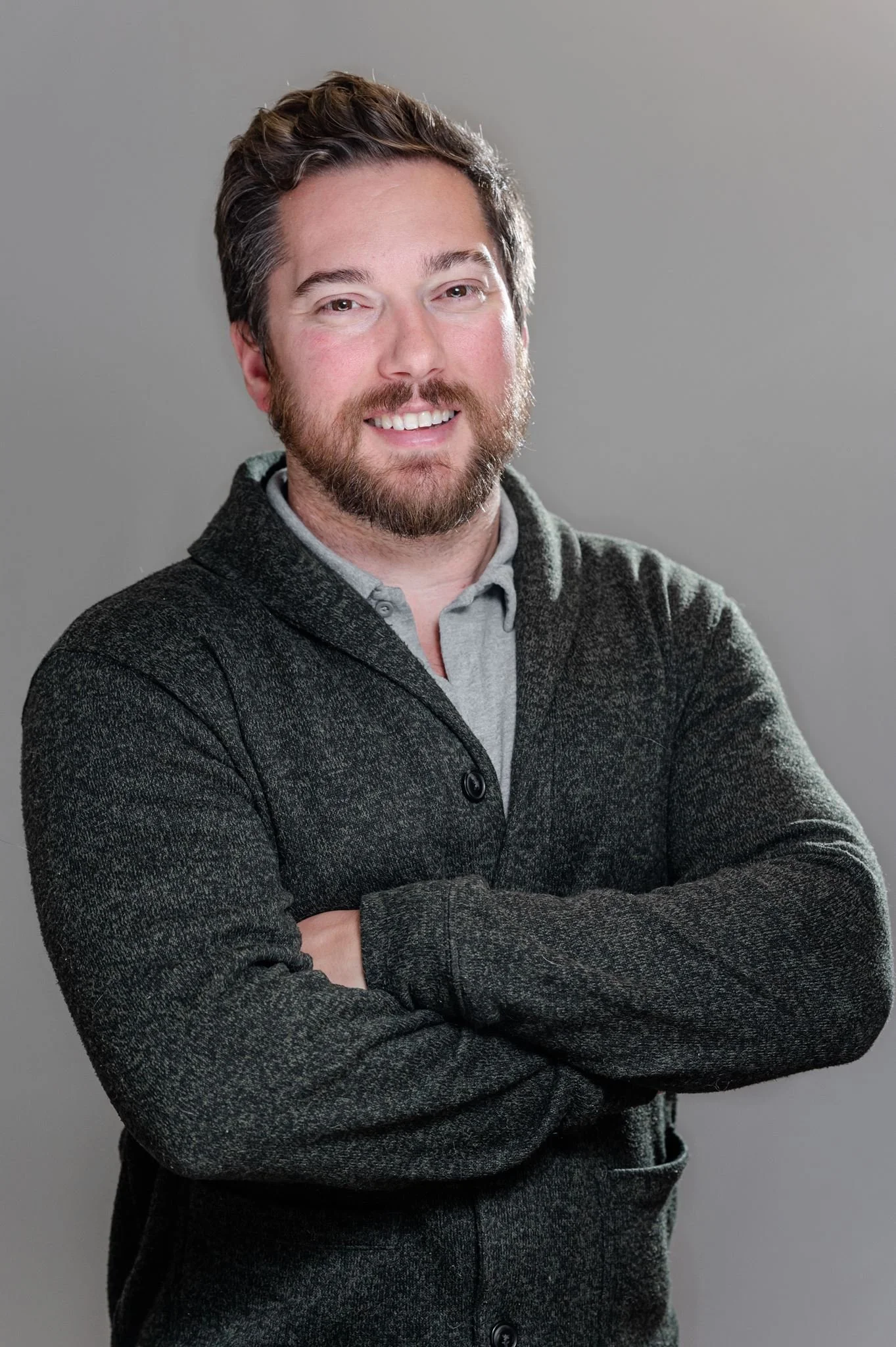 A man with brown hair, beard, and mustache smiling, wearing a gray collared shirt under a dark gray textured cardigan, standing with arms crossed against a plain gray background.