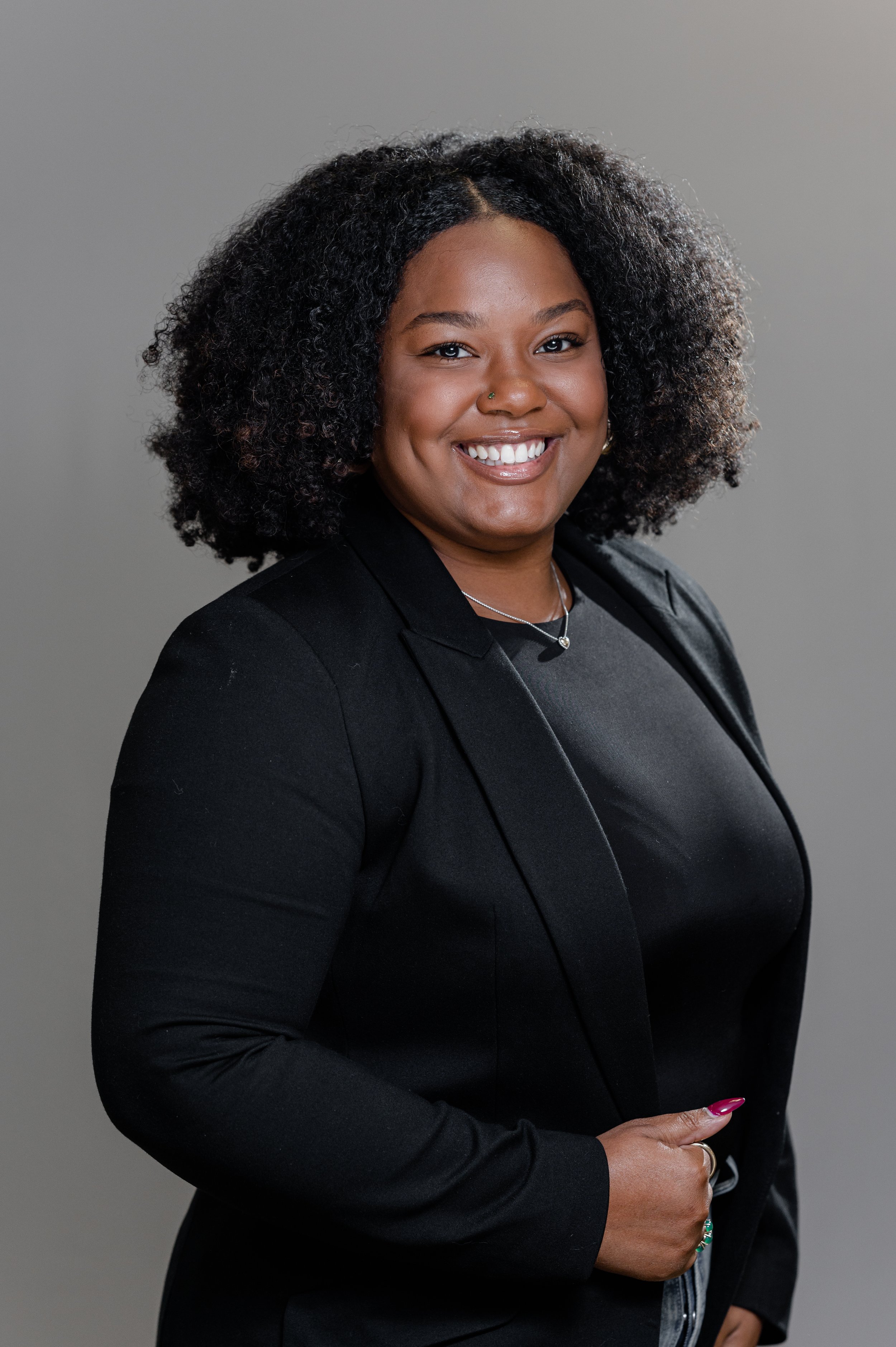 A professional headshot of a black woman with curly black hair, wearing a black blazer and silver jewelry, smiling with a gray background.