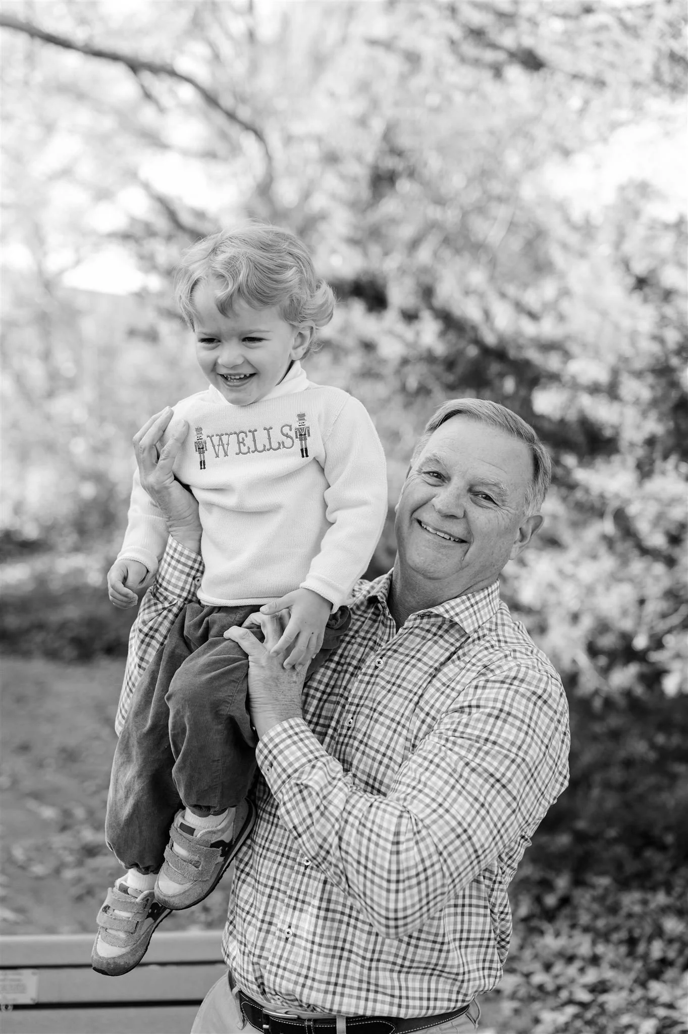 An elderly man is holding a young boy on his shoulder outside in a park with trees in the background. The boy is smiling and wearing a sweatshirt that says "WELLS." The man is smiling and wearing a checkered shirt.