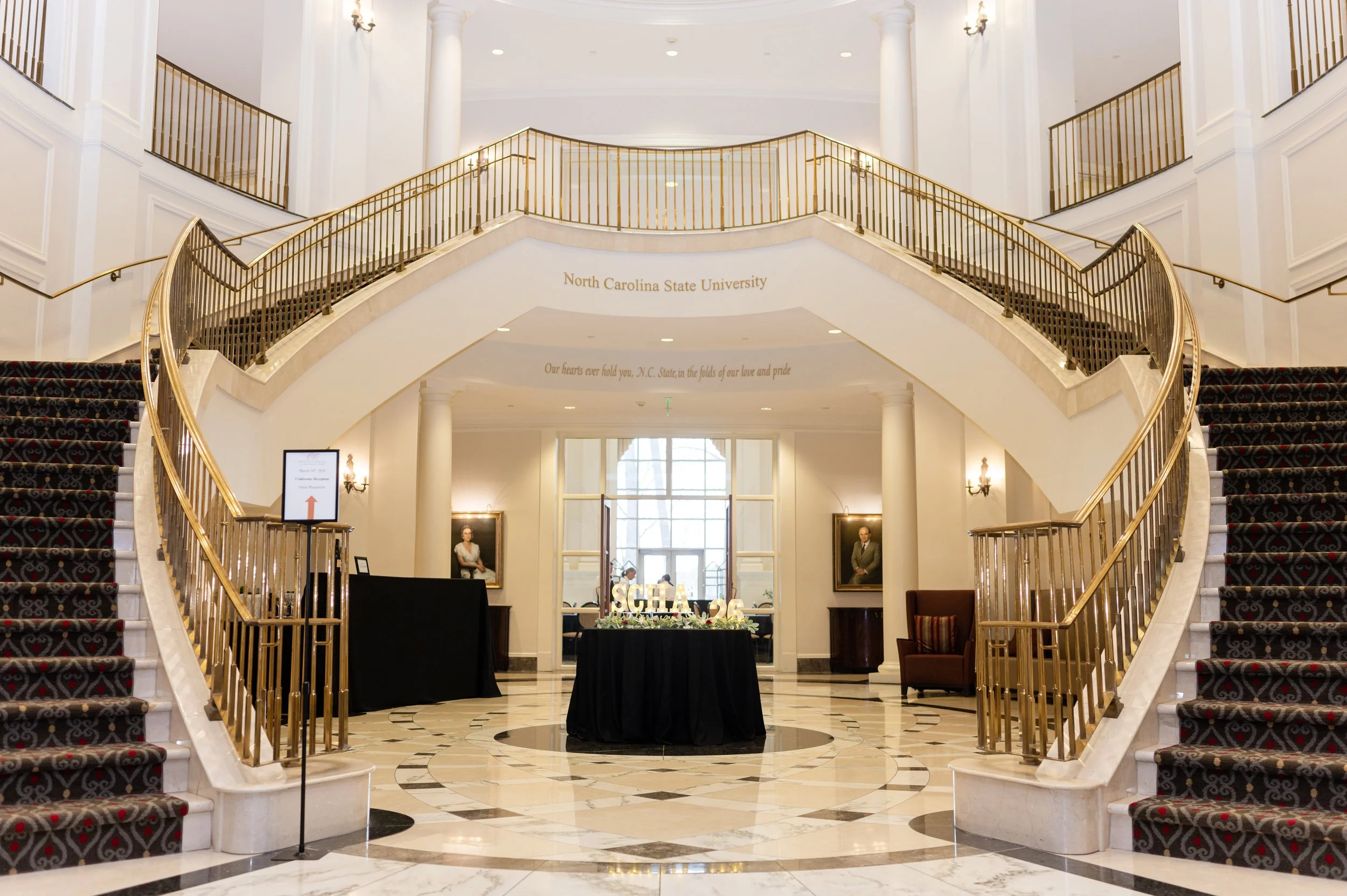 Inside a grand building with a double staircase, gold railings, and white columns, featuring a sign for North Carolina State University and a table with flowers and a sign that reads 'SCHA 2026'.