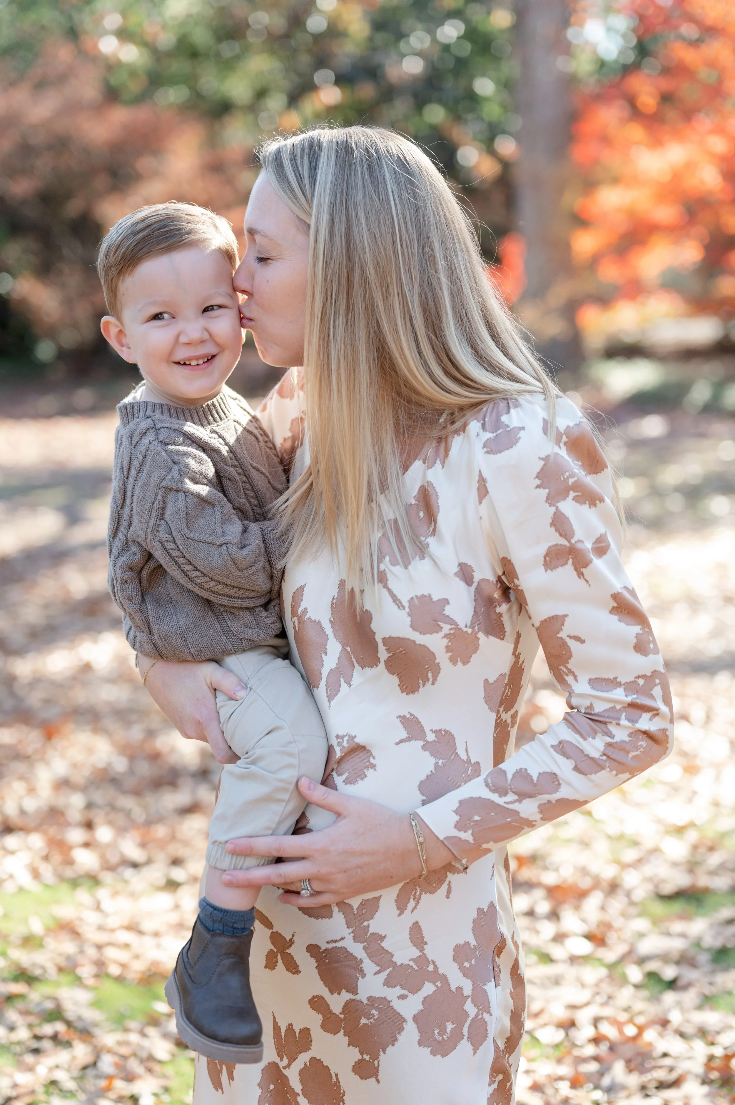 A woman holding a young boy outdoors during autumn, with colorful fall leaves in the background. The woman is kissing the boy on the cheek, and both are smiling.