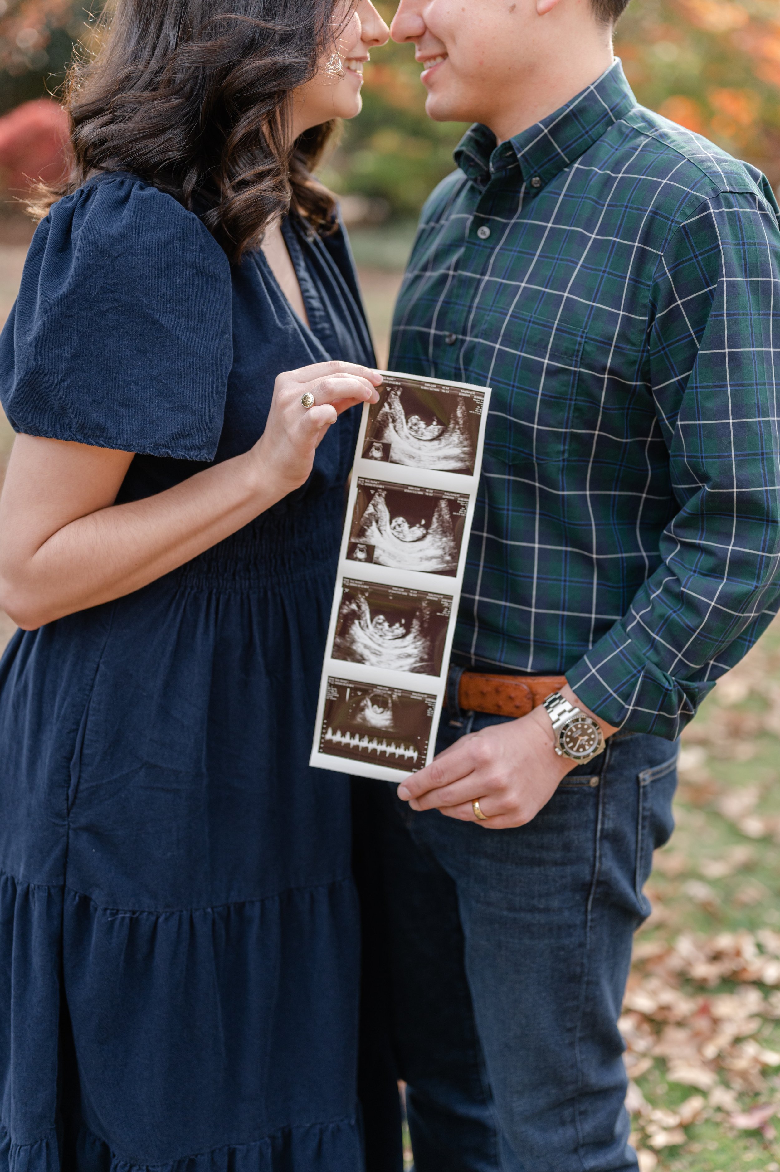 A couple standing close together outdoors, holding an ultrasound image of a baby, smiling and touching foreheads, with autumn trees in the background.
