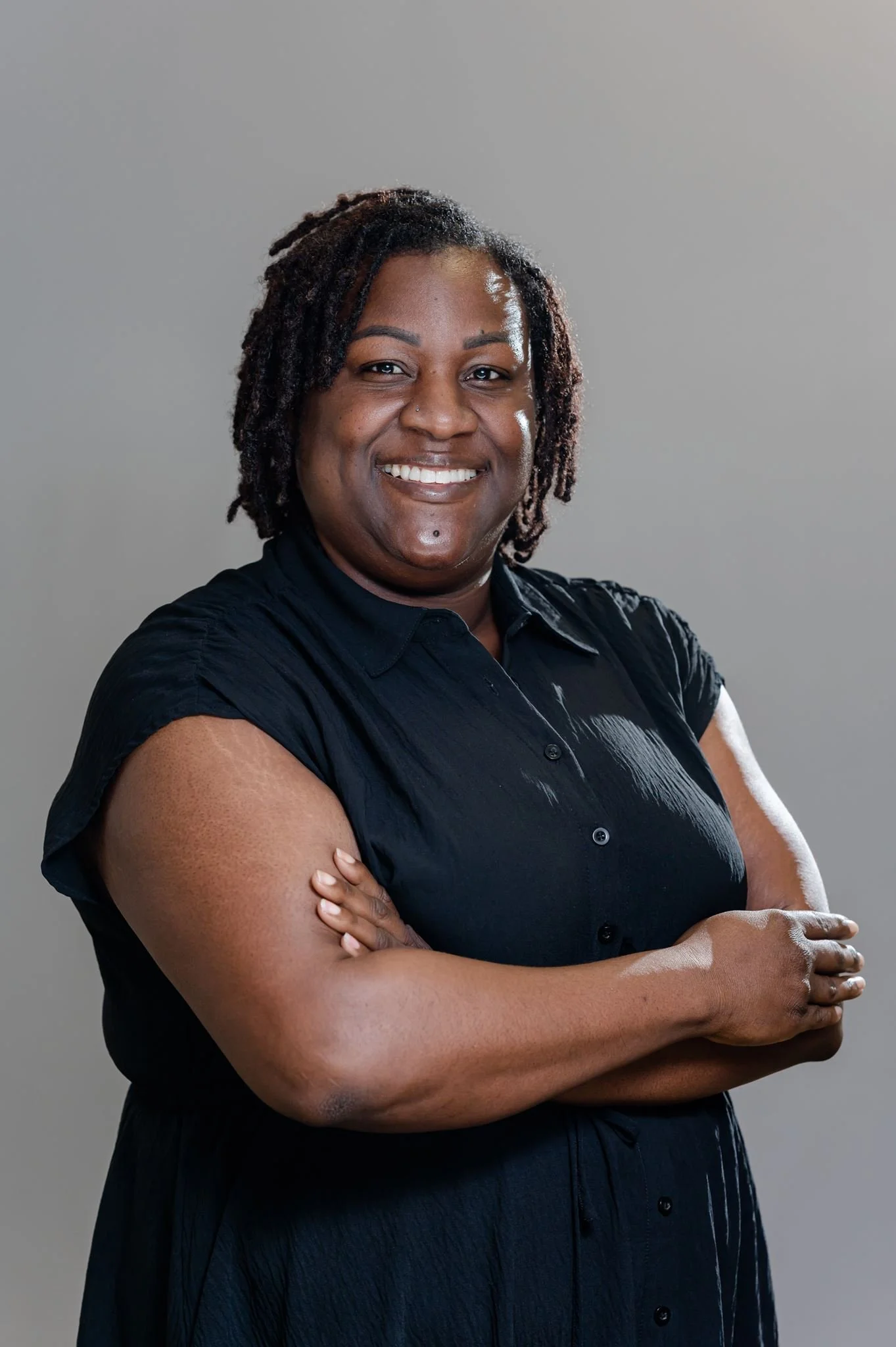 A professional headshot of a smiling black woman with medium brown skin and dreadlocks, wearing a black shirt with short sleeves, standing with her arms crossed against a plain grey background.