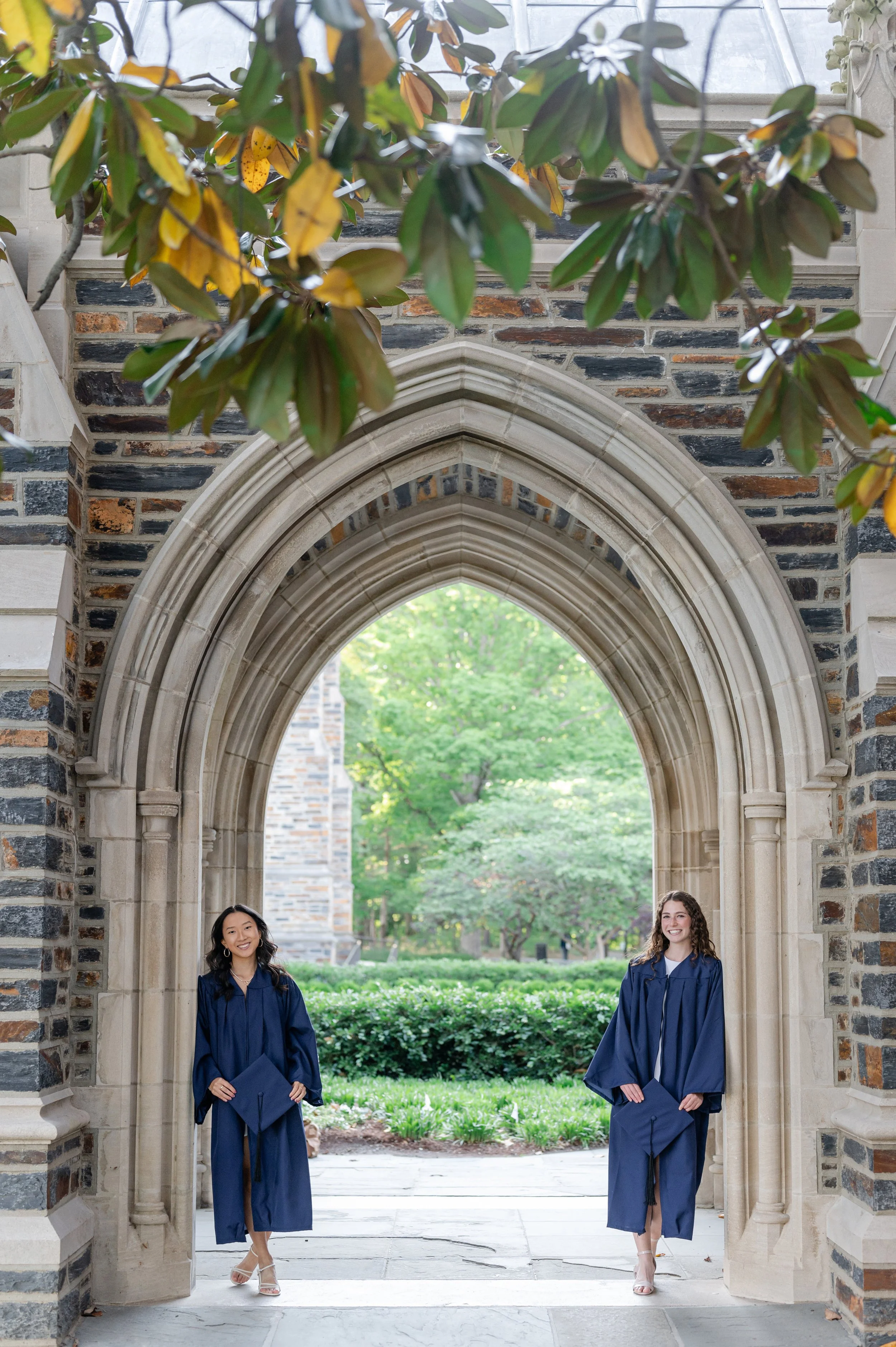 Two women in blue graduation gowns and holding diplomas standing under an arched stone structure with trees in the background.