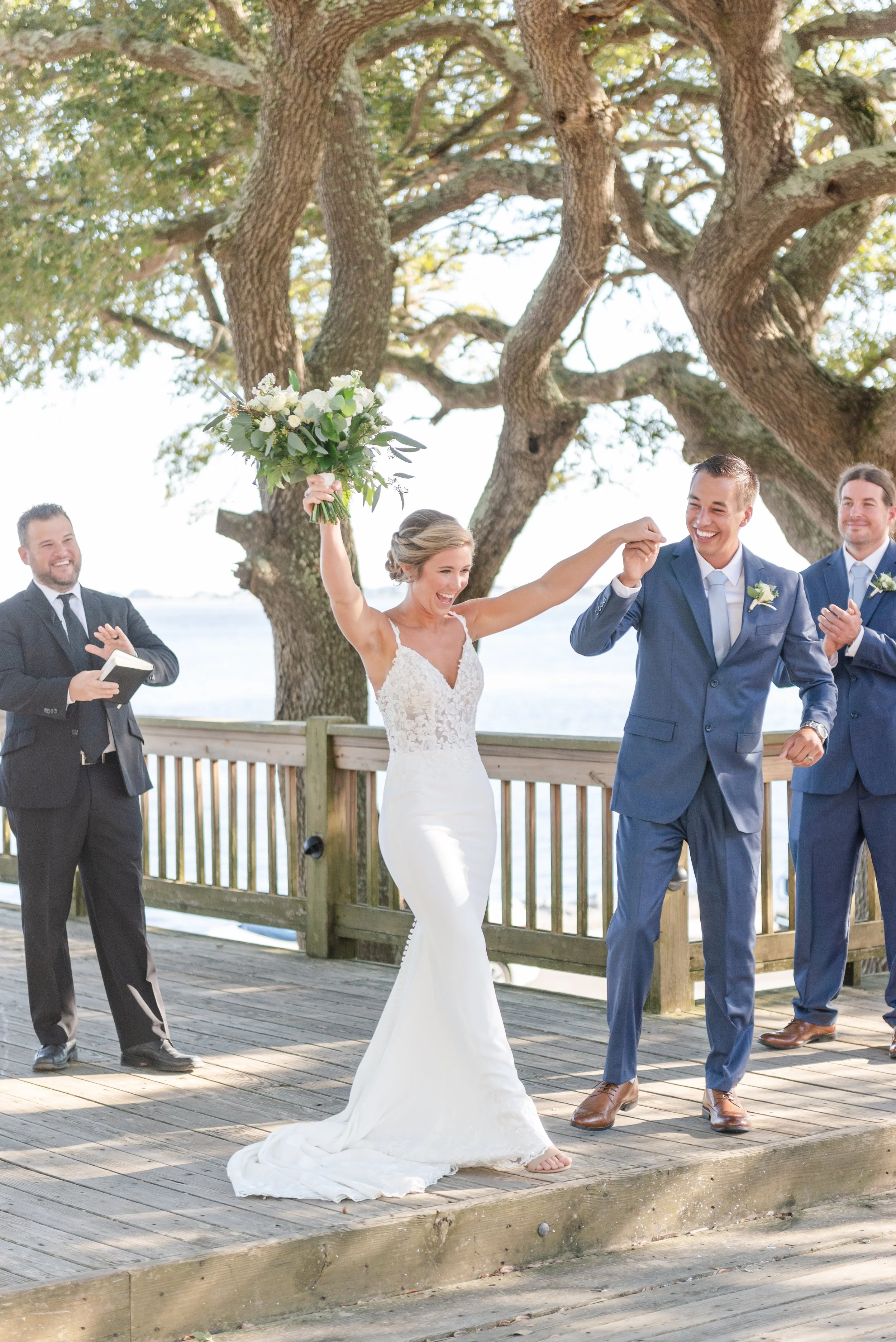 Bride and groom celebrating with raised hands on a wooden deck, surrounded by wedding officiant and groomsmen, with trees and water in the background. The bride holds a bouquet.