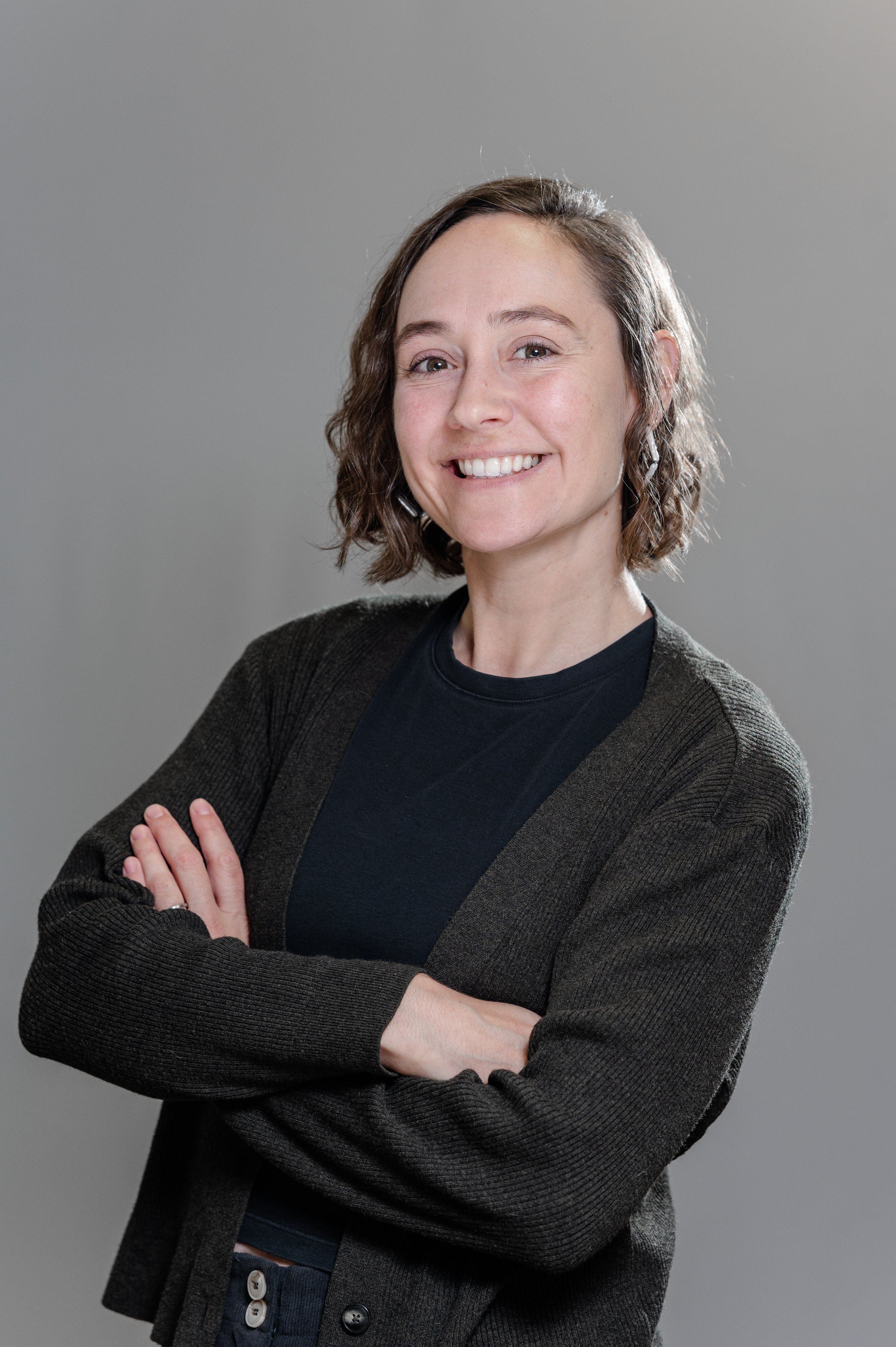 A woman with short wavy brown hair, smiling with arms crossed, wearing a black shirt and dark gray cardigan against a plain gray background.