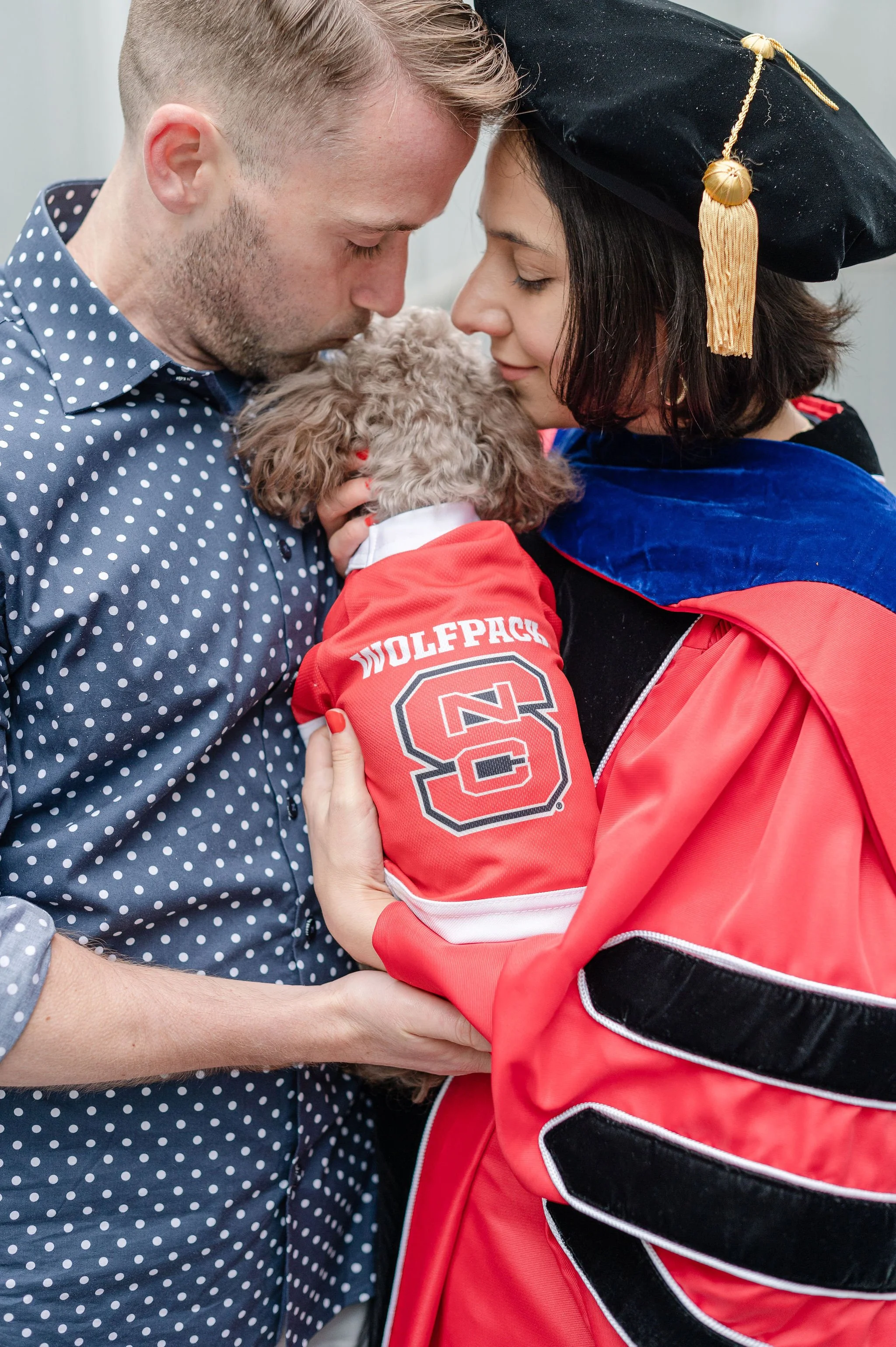 A person in a graduation cap and gown holding a small dog in a red college hoodie, with face close to two other people, all sharing a tender moment.