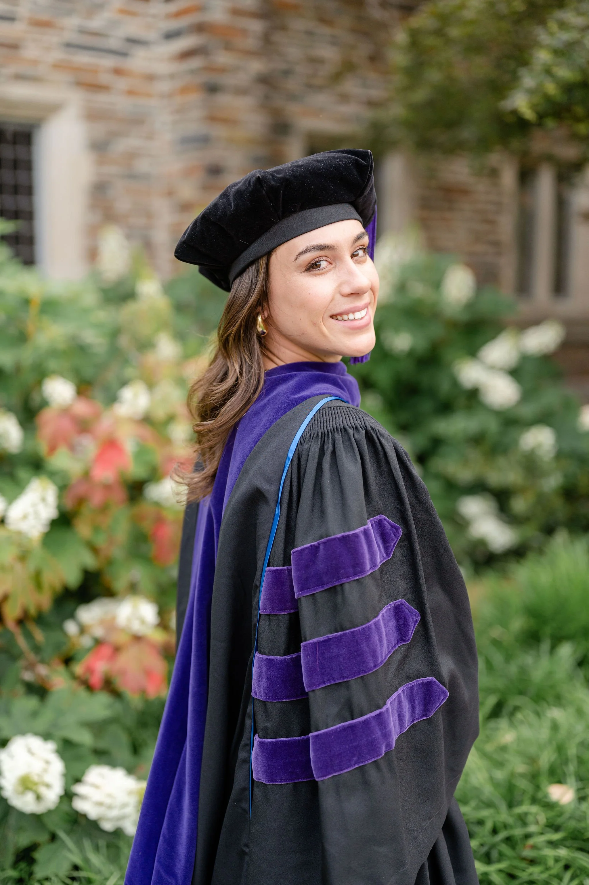 A young woman in a graduation gown and cap, smiling and looking back, standing outdoors with a background of flowers and a brick building.