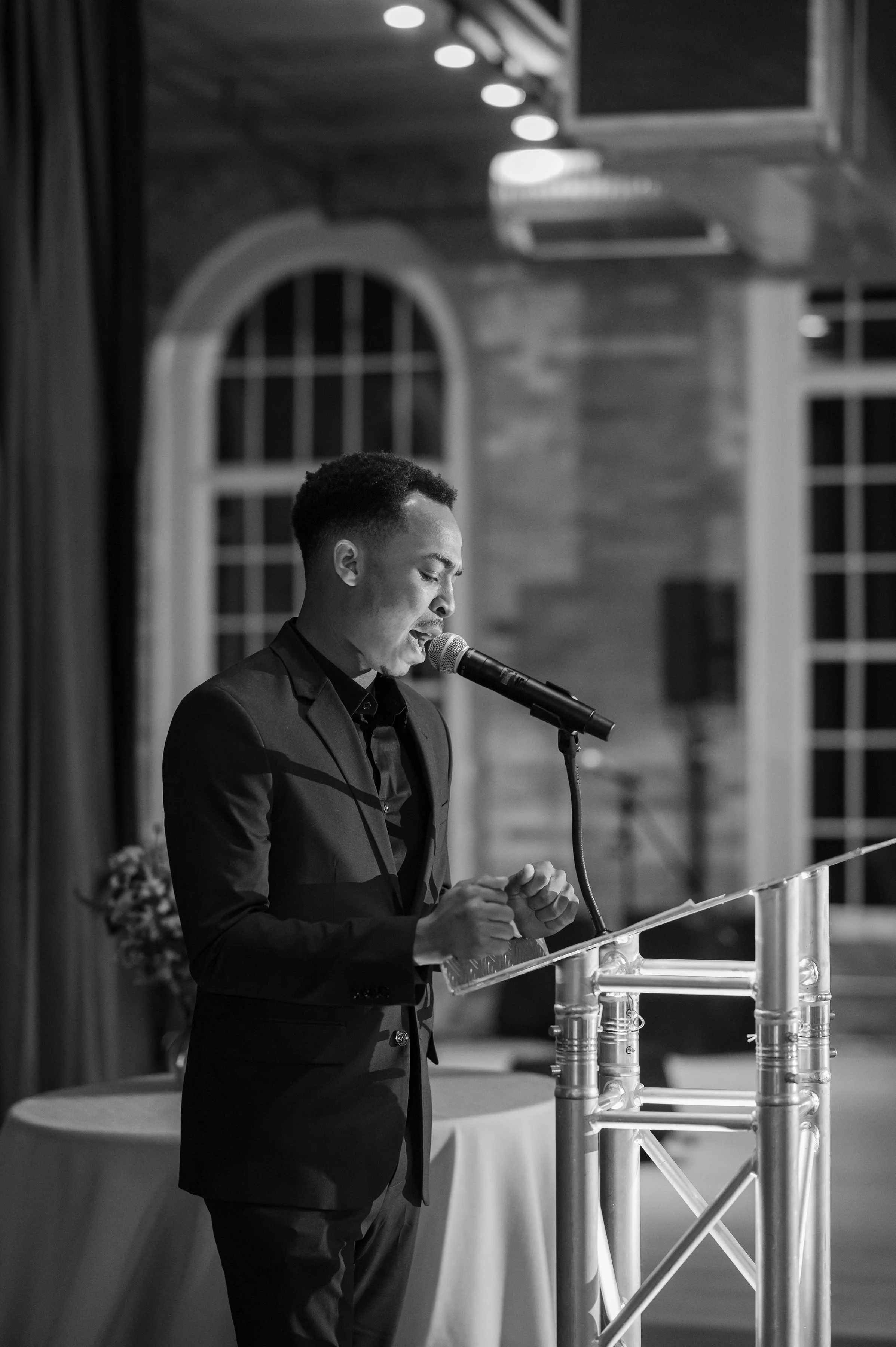 A man in a dark suit is standing at a transparent podium, speaking into a microphone, in a room with large arched windows and wooden walls, in black and white.
