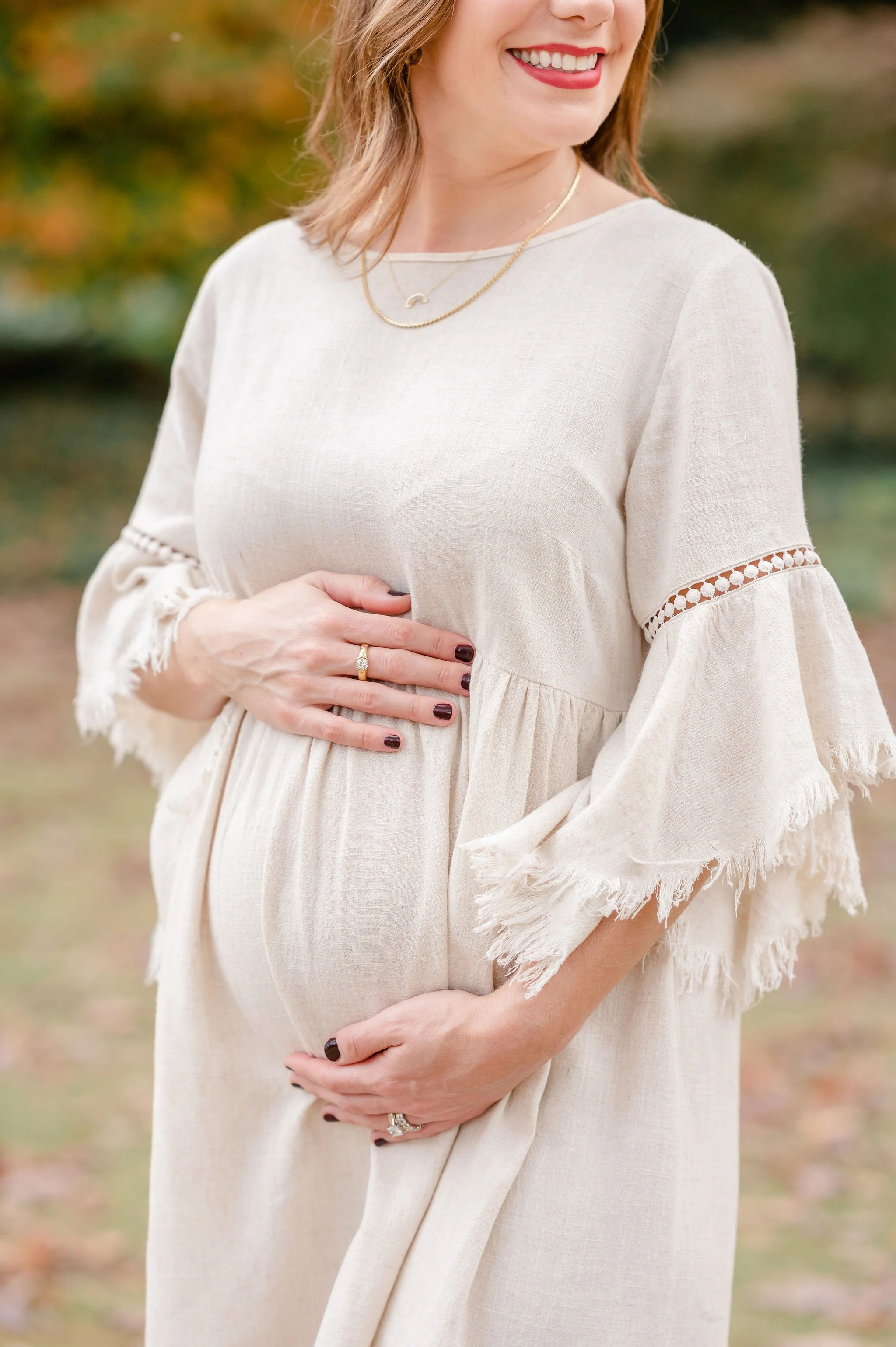 Close-up of a woman smiling outdoors, wearing a beige dress with lace details, gold jewelry, and holding her hand over her stomach.